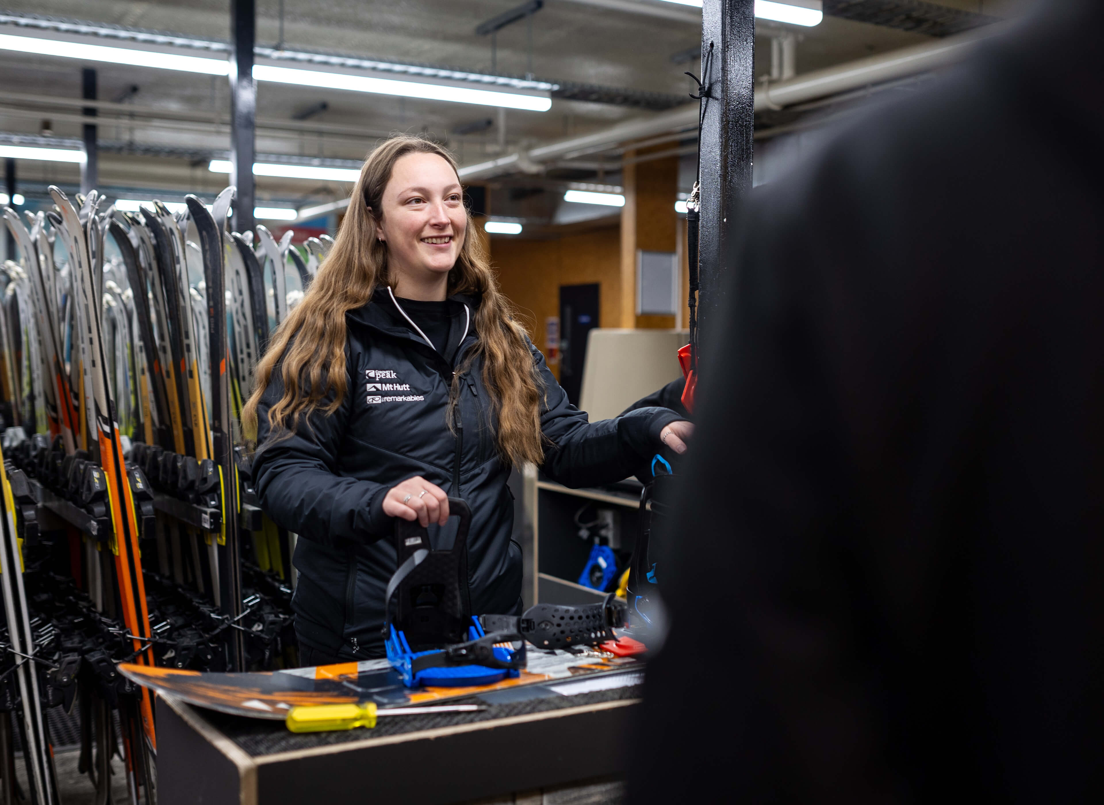Smiling woman assisting customer at ski rental counter with skis in background.