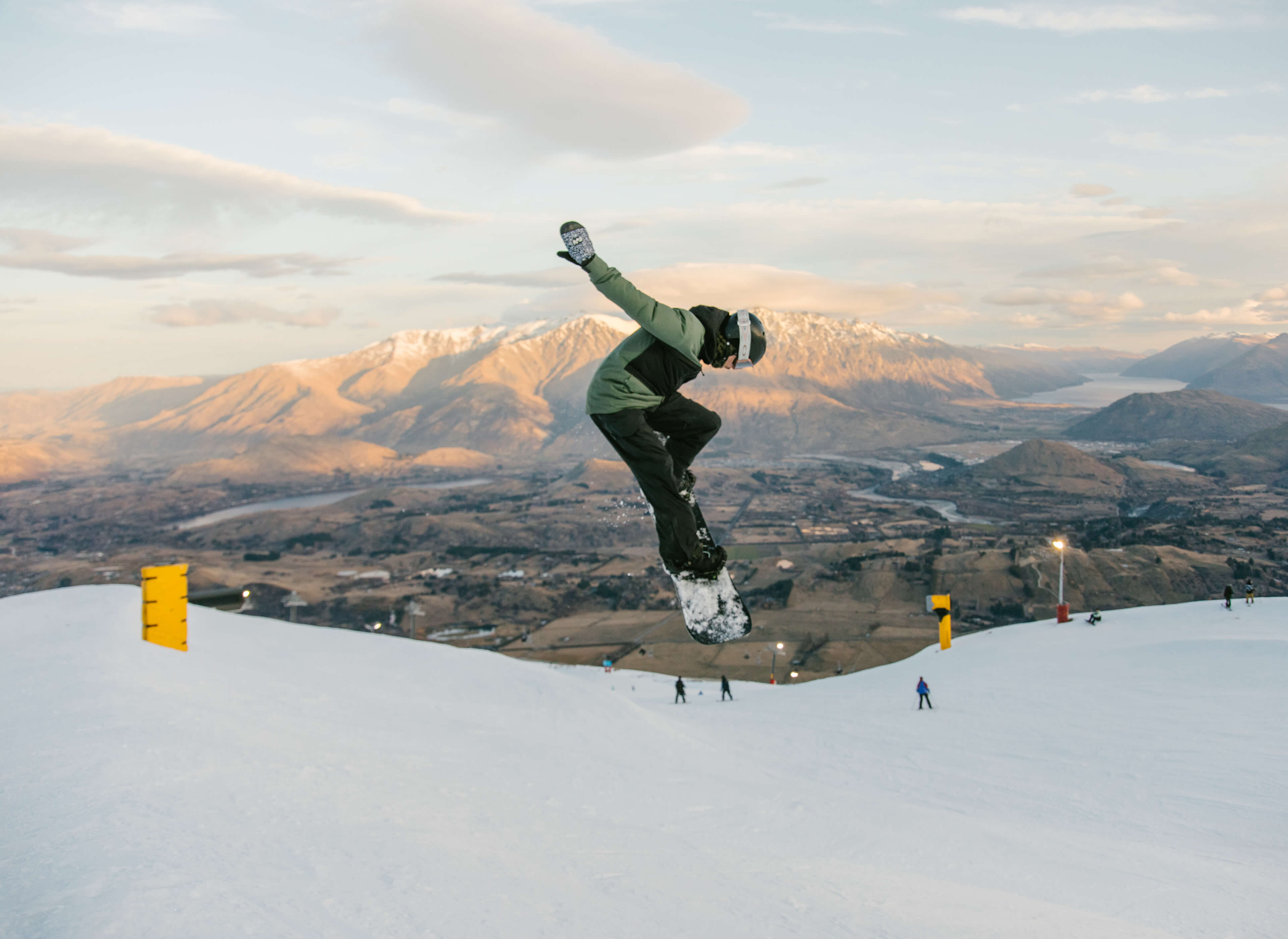 Snowboarder in green and black gear jumps on a snowy slope with mountainous valley in the background.