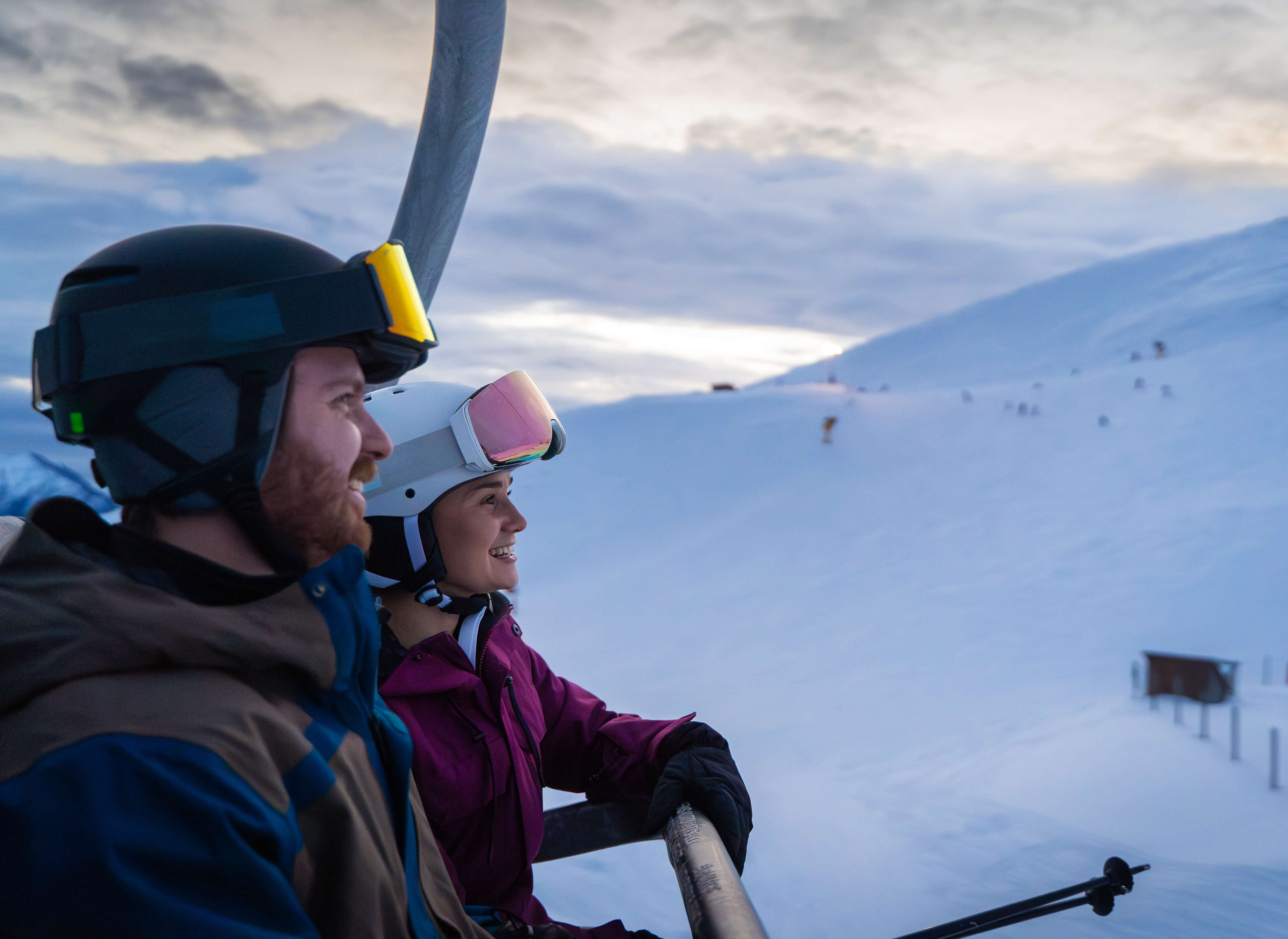 Man and woman in ski gear riding a chairlift above snowy slopes near sunset.