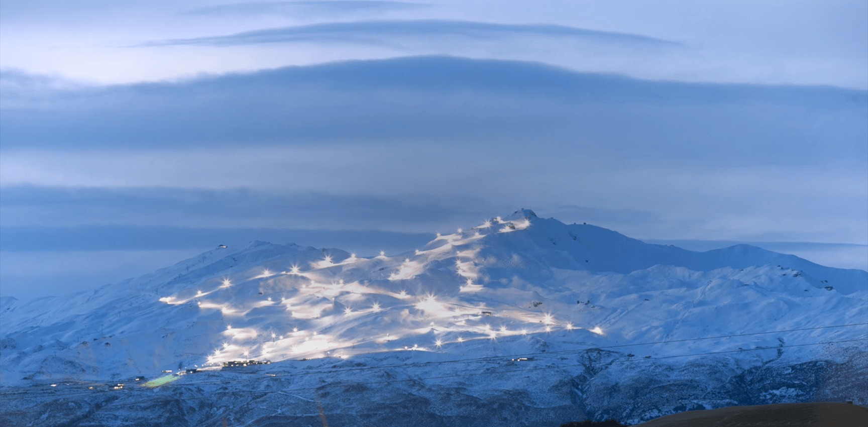 Snow-covered mountain ski slopes illuminated with lights under a blue twilight sky.