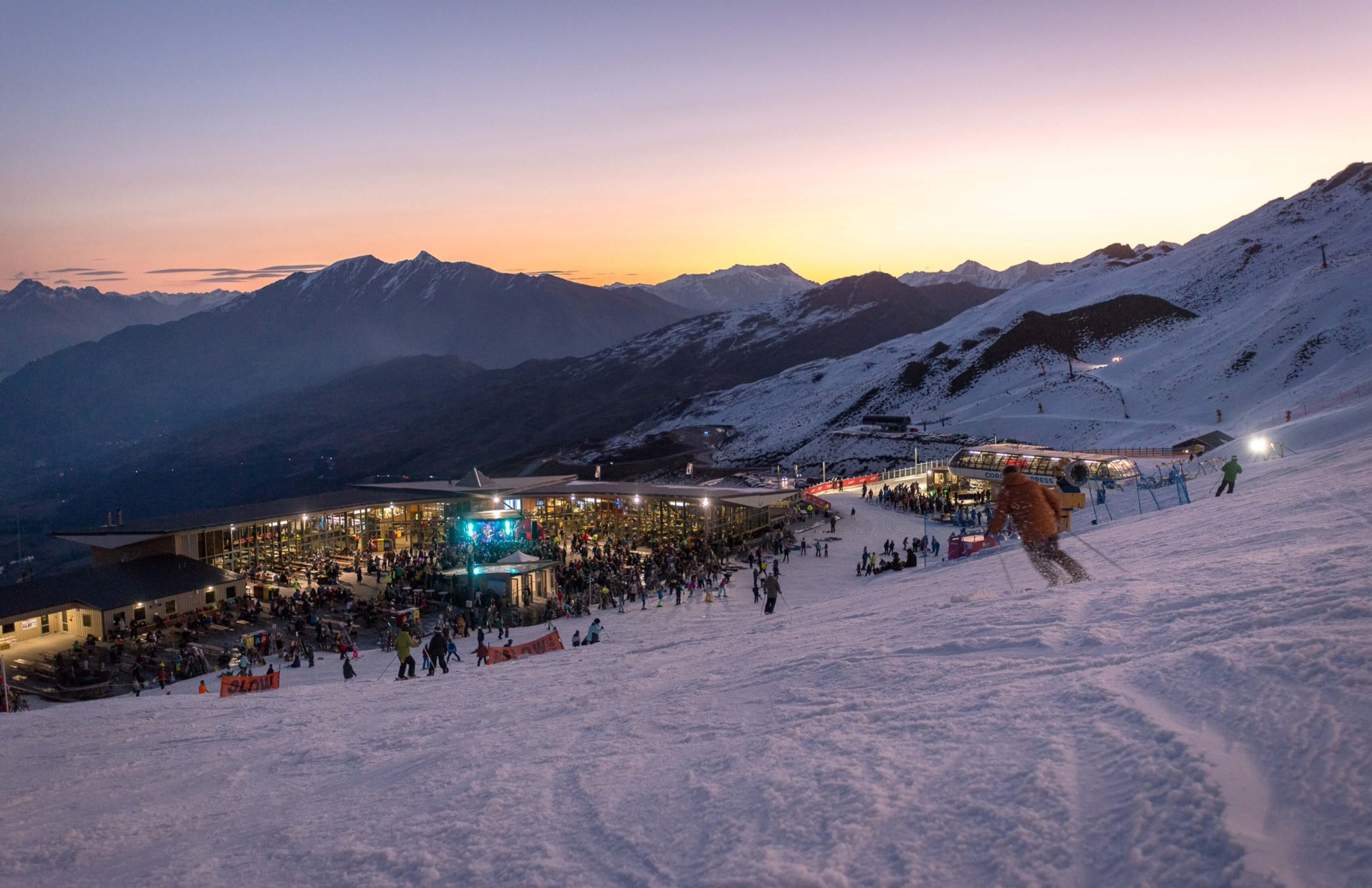 Skiers on snowy slope near lit lodge at sunset with mountain range in background.
