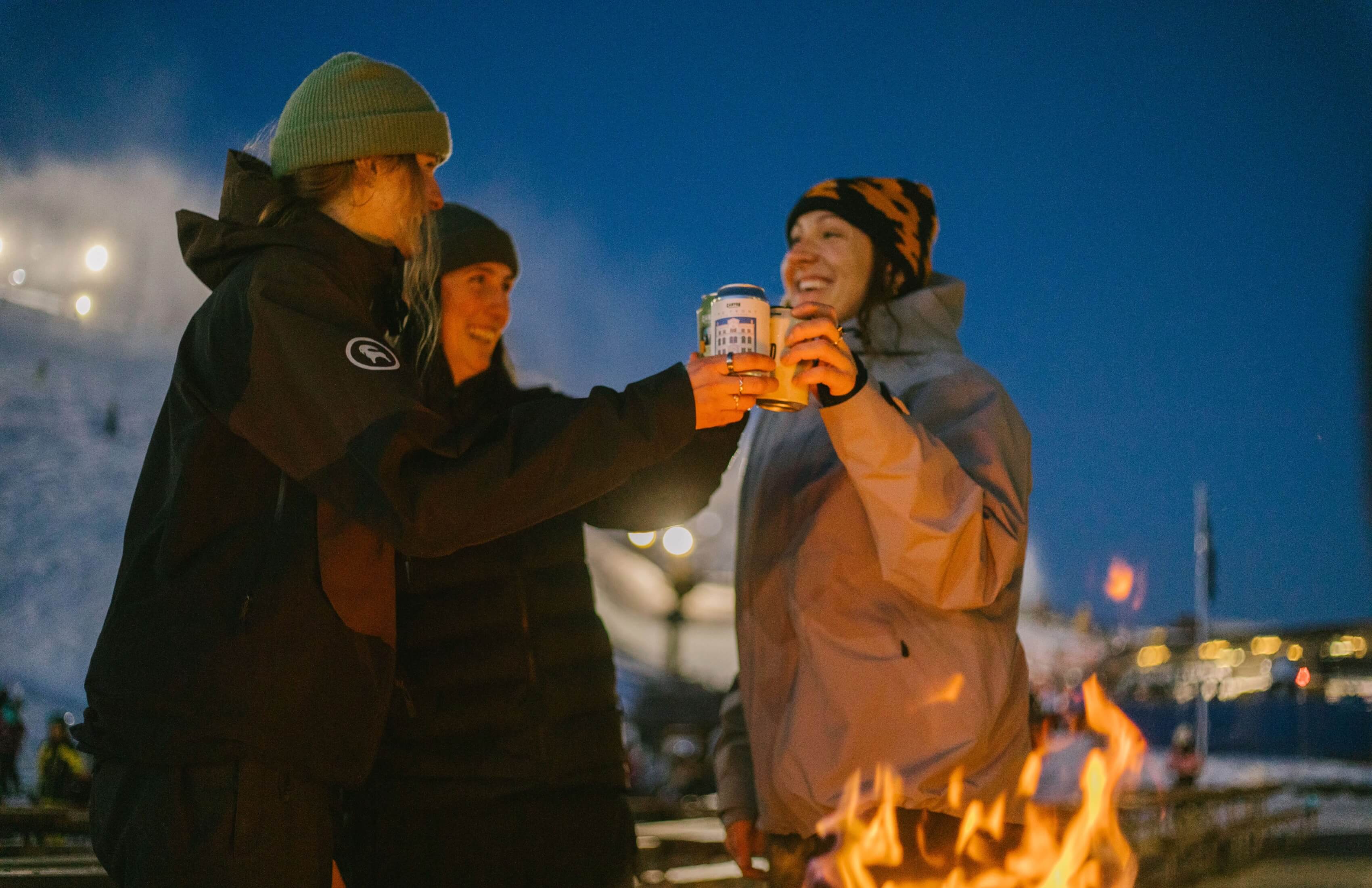 Three friends in winter clothes toasting with cans near a fire at night.