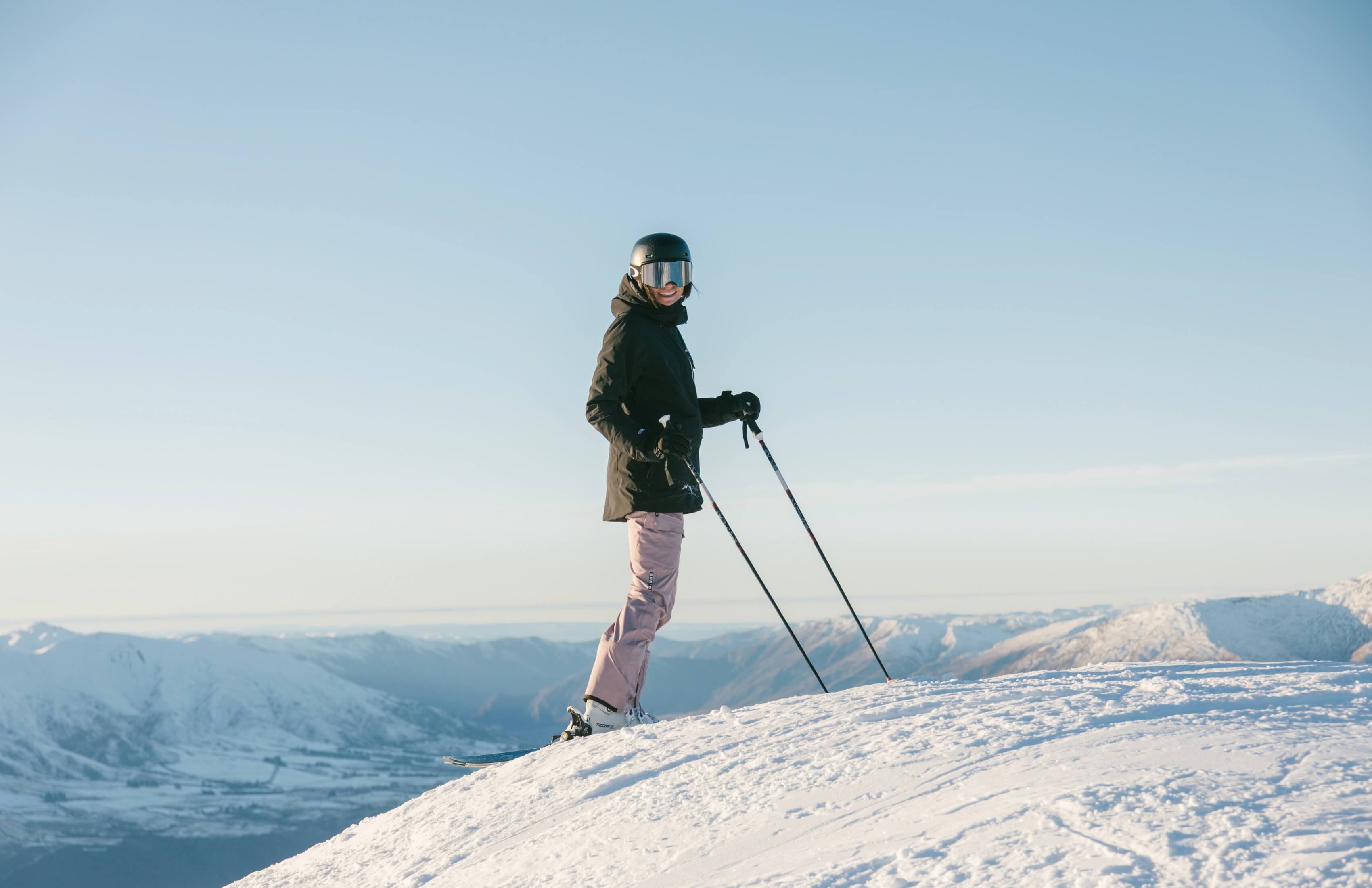 Smiling skier in black jacket and helmet standing on snowy slope with mountain views under clear sky.