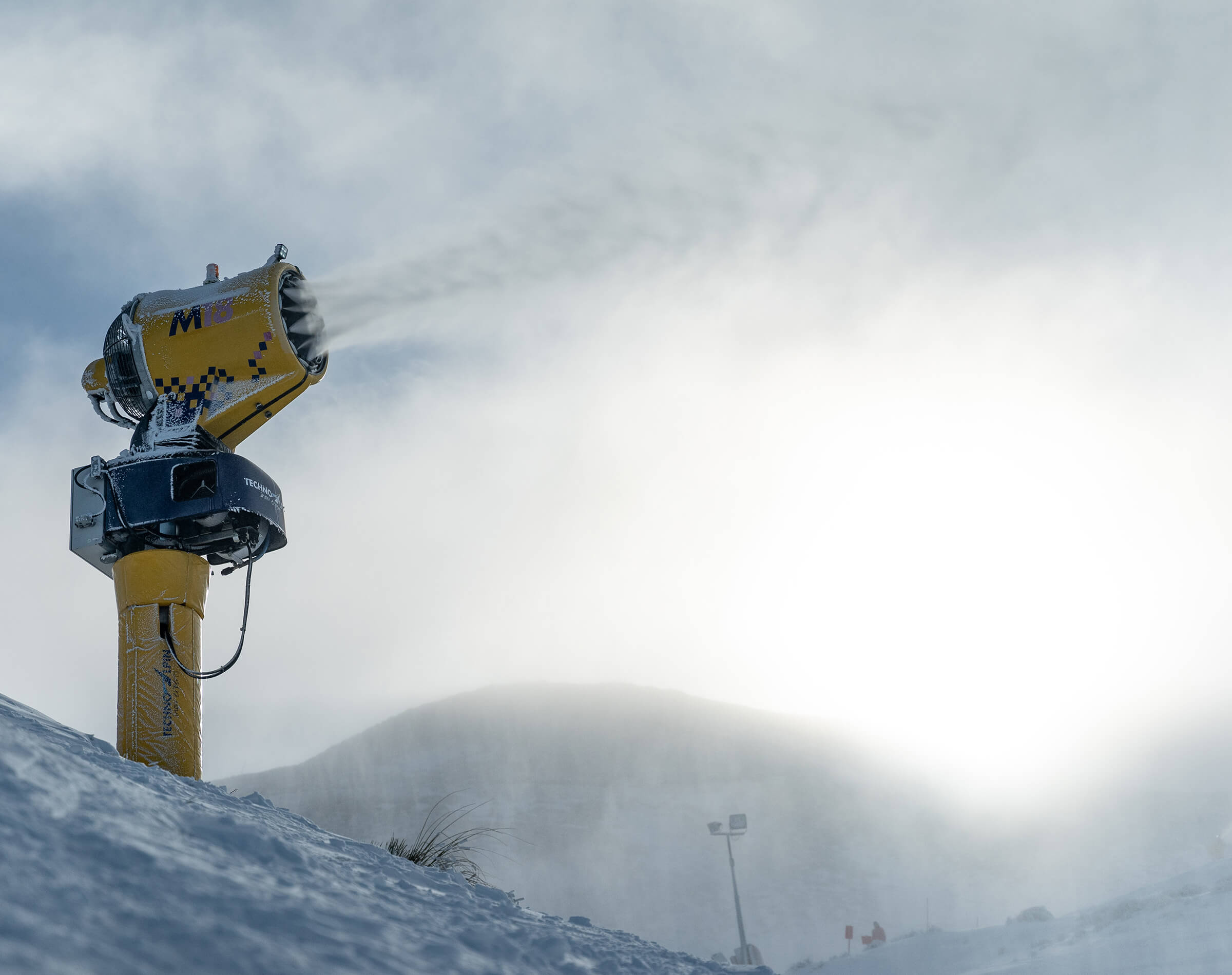 Yellow snow-making machine spraying snow on a snowy mountain slope in bright daylight.