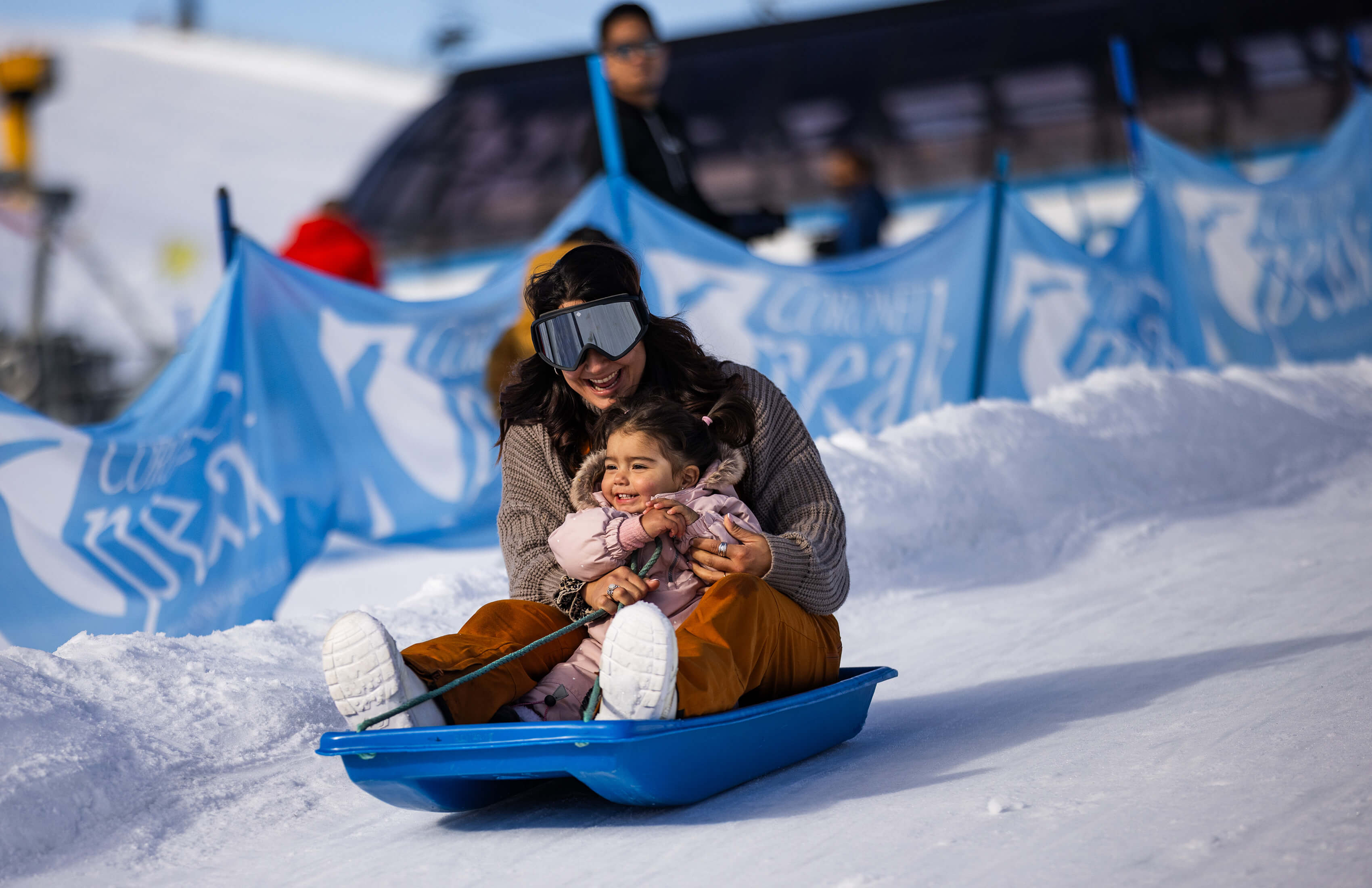 Smiling woman and child sledding down a snowy hill on a blue sled.