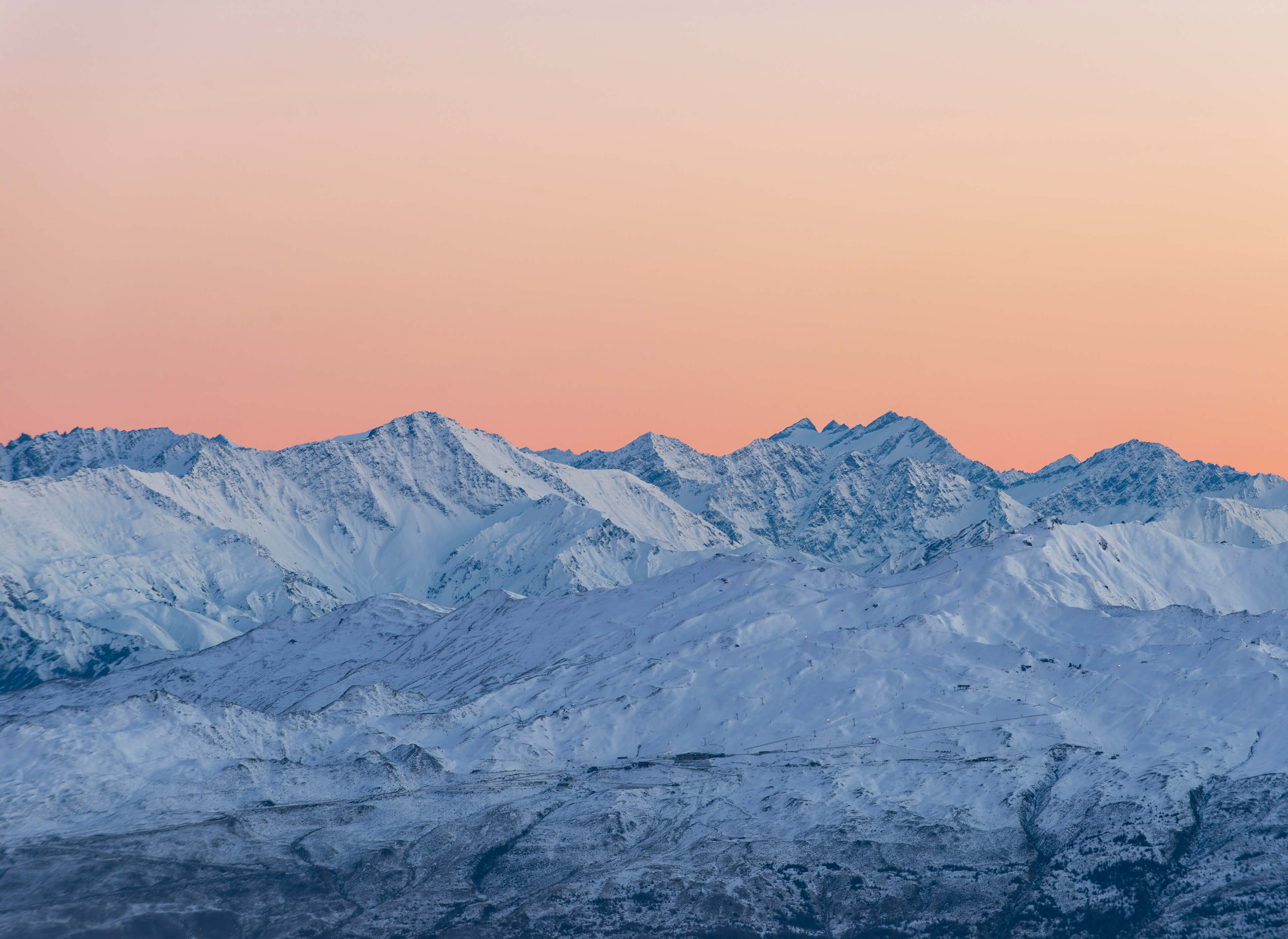 Snow-covered mountain range under a clear orange-pink sky at sunset or sunrise.