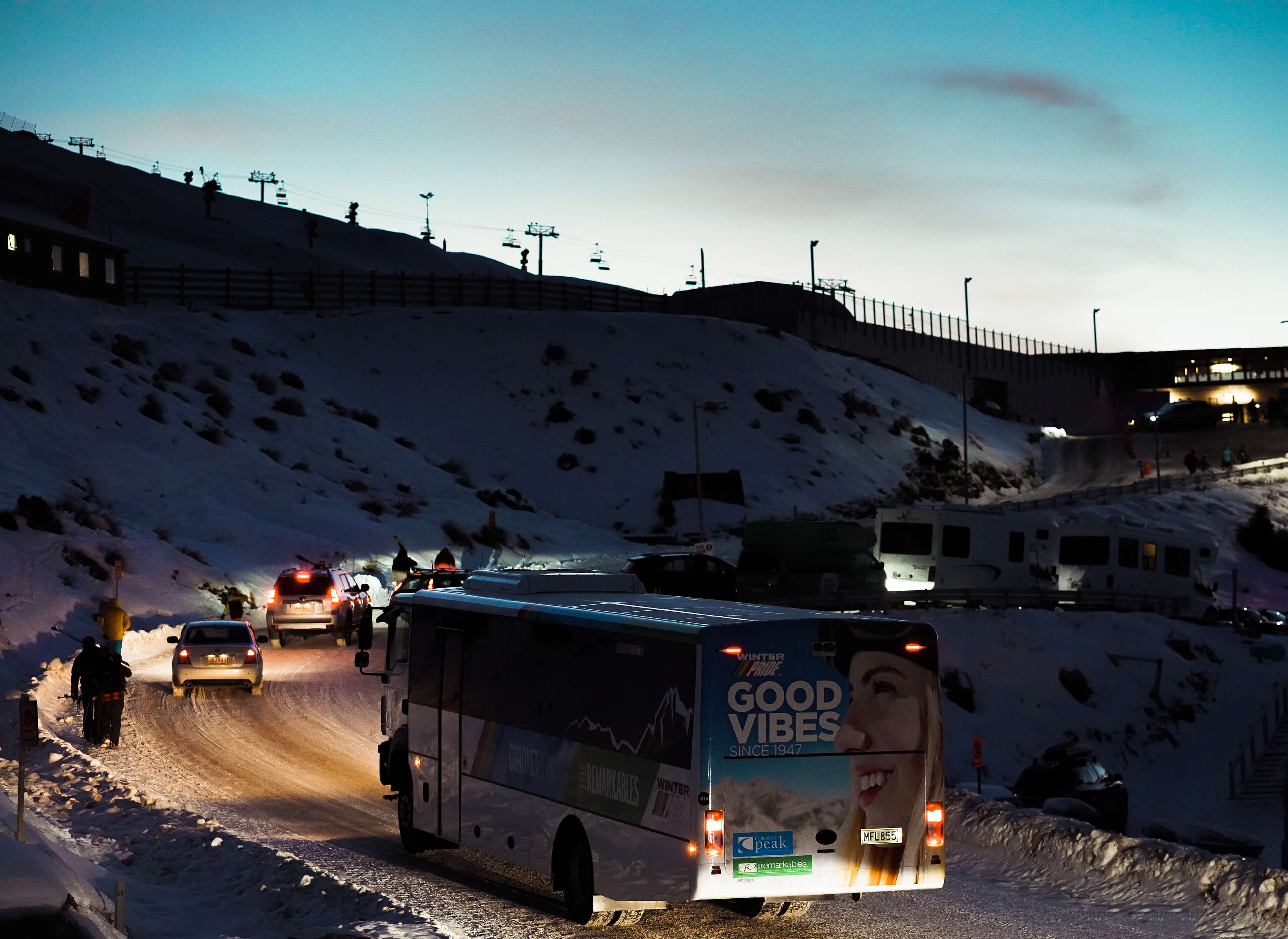Bus with 'Good Vibes' text driving on snowy mountain road at dusk with cars and ski lifts nearby.