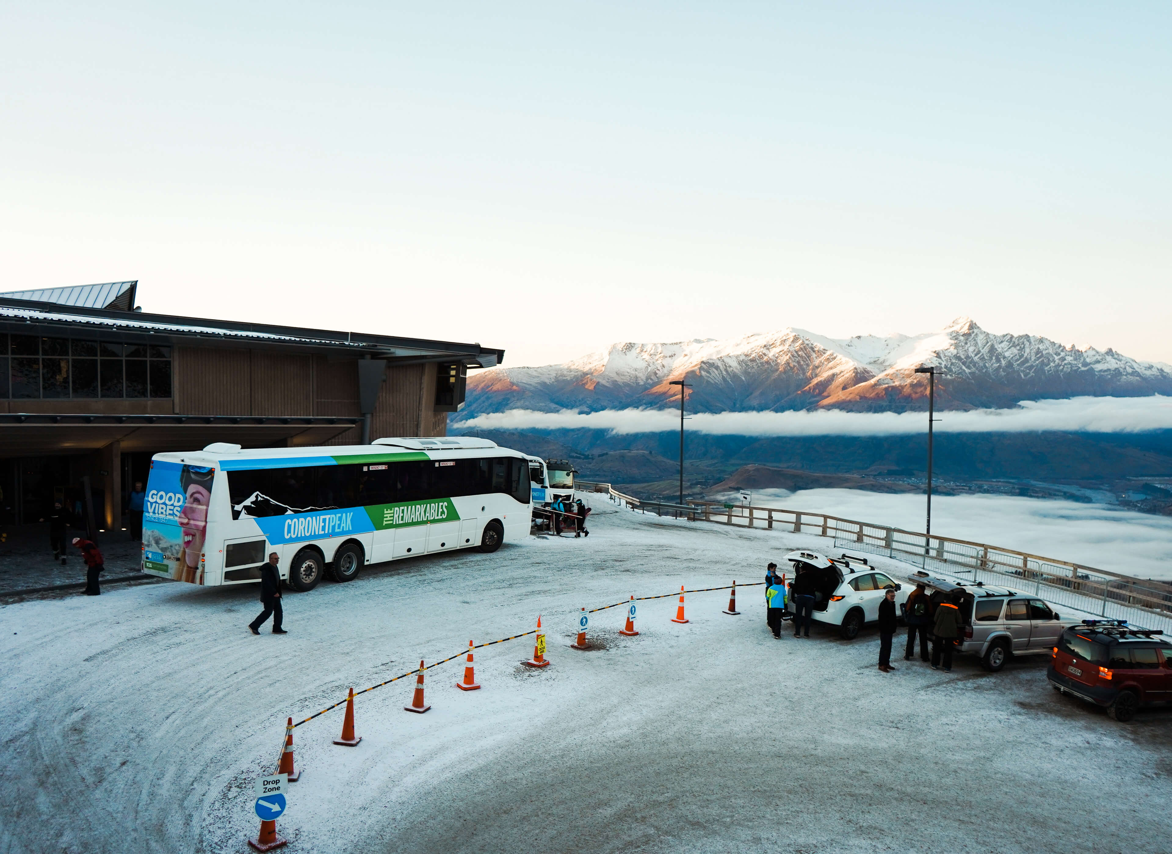 Bus and cars parked at snowy mountain drop zone with people and snow-capped peaks in the background.