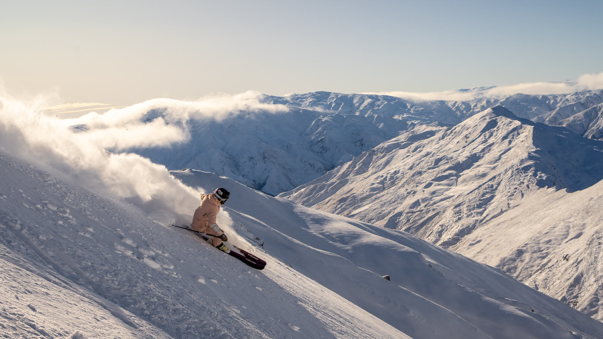 Skier in beige jacket carving down snowy slope with mountain range in the background.