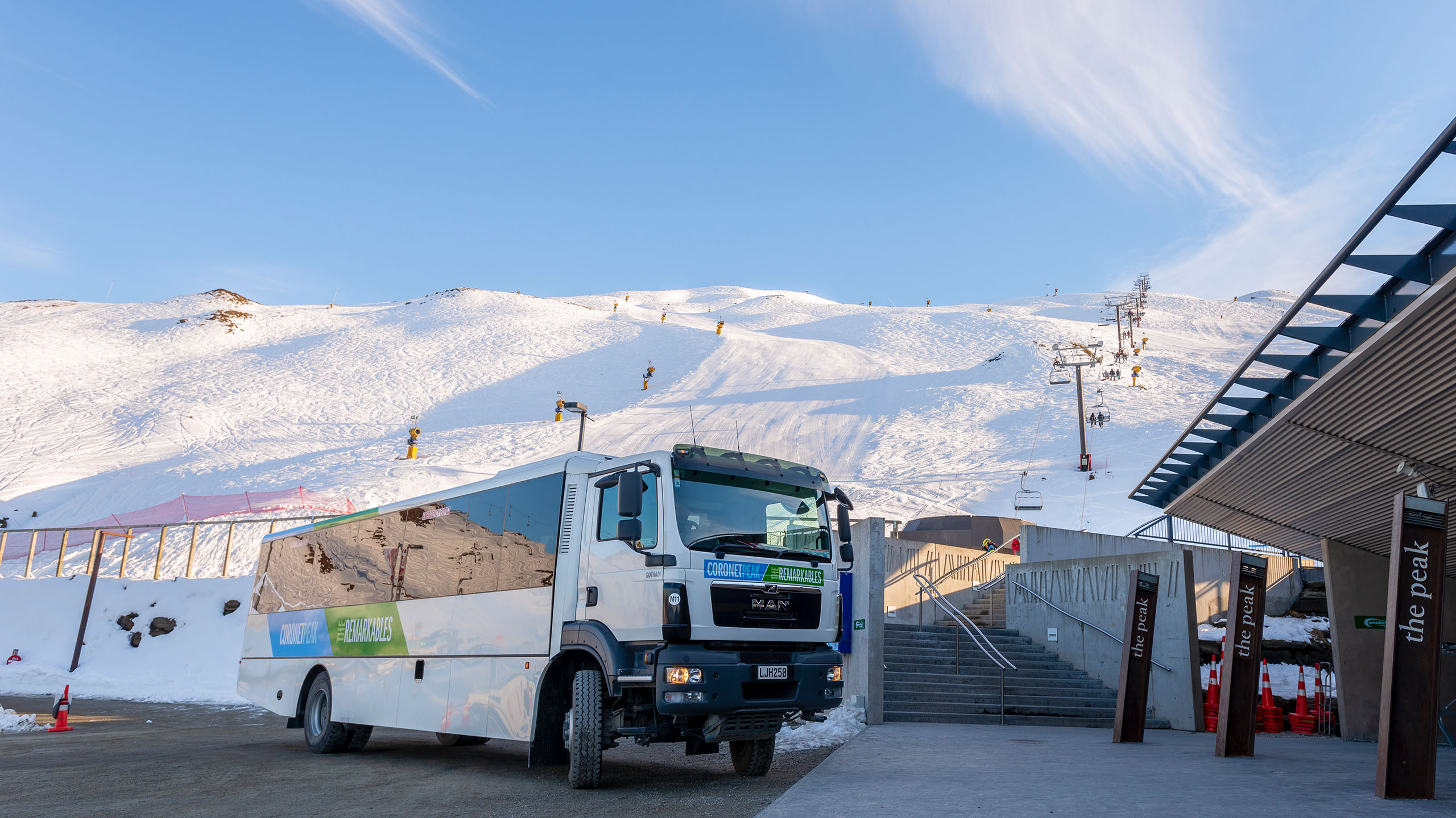 White bus parked near ski lift station with snow-covered hills and clear blue sky in background.