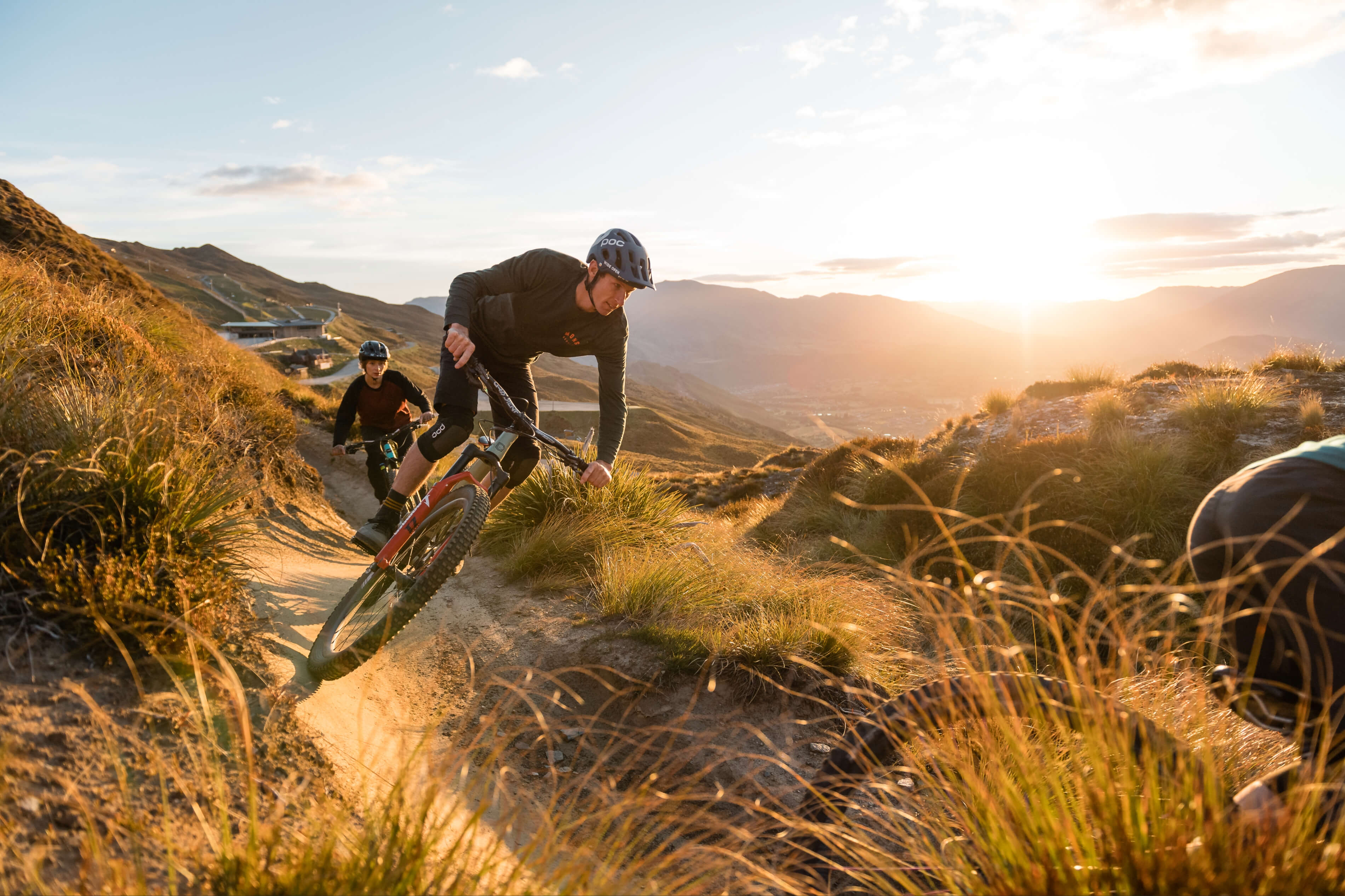 Two cyclists ride mountain bikes on a dirt trail at sunset in a hilly landscape.