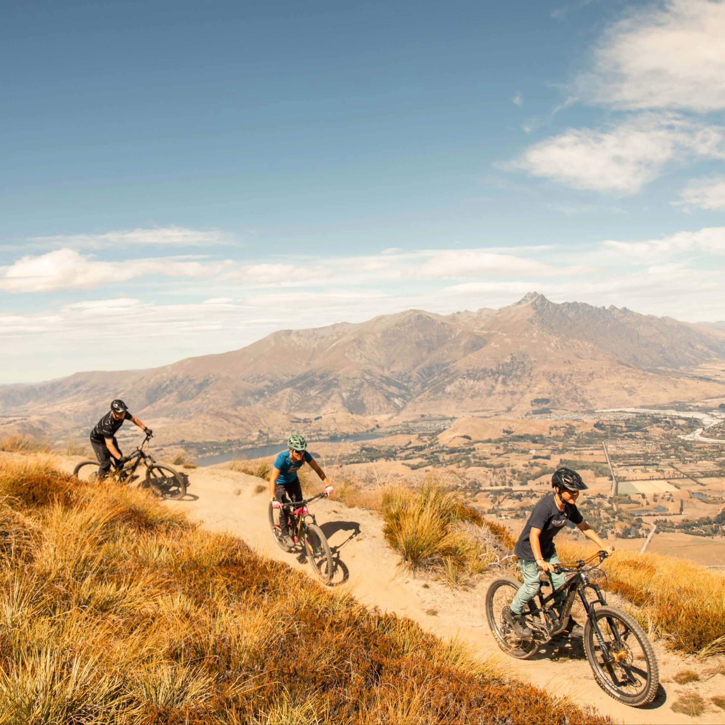 Three mountain bikers riding a dirt trail through dry grass with mountains and valley in background.