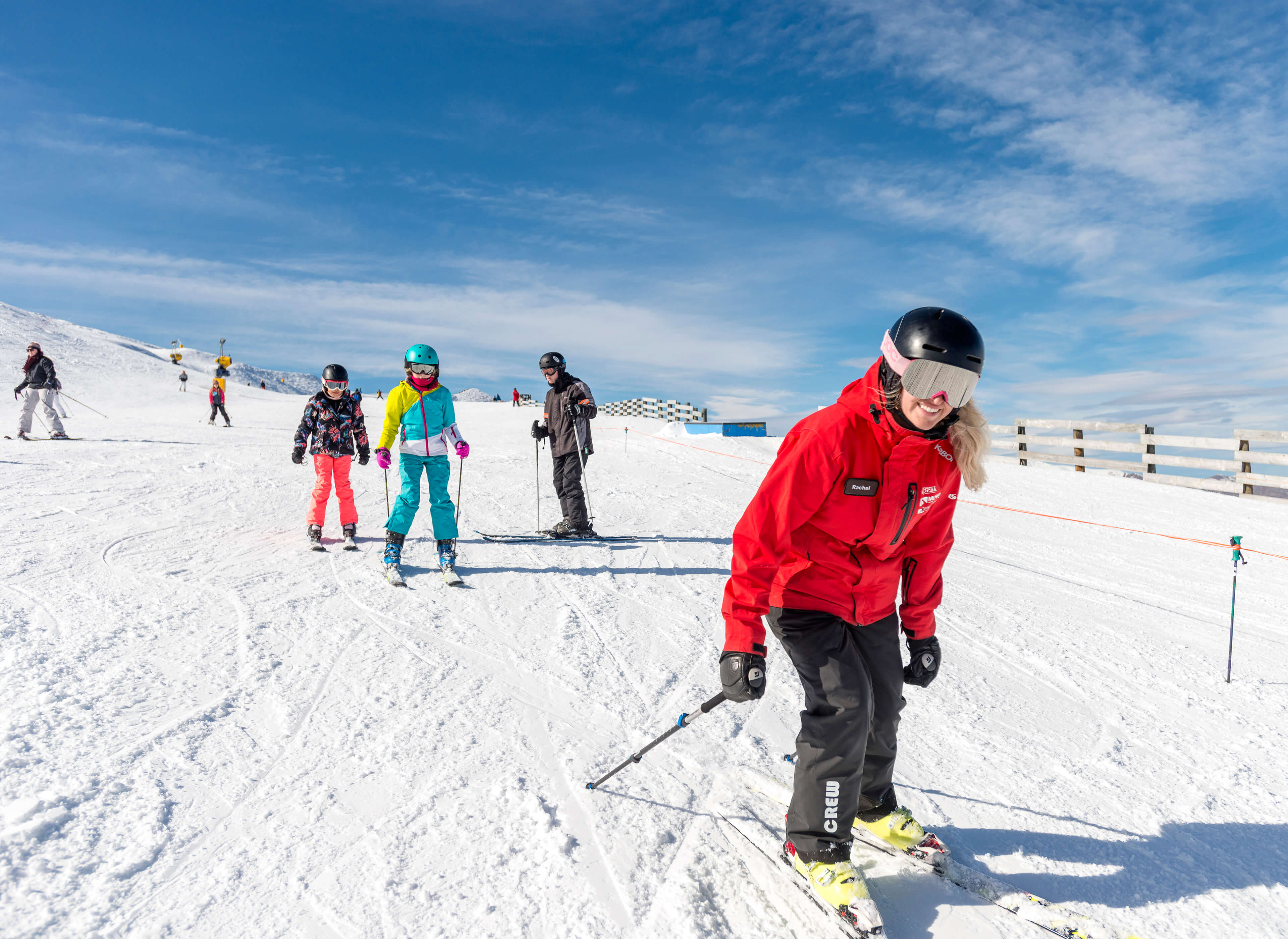 Ski instructor in red jacket teaching three children skiing on a snowy slope under blue sky.
