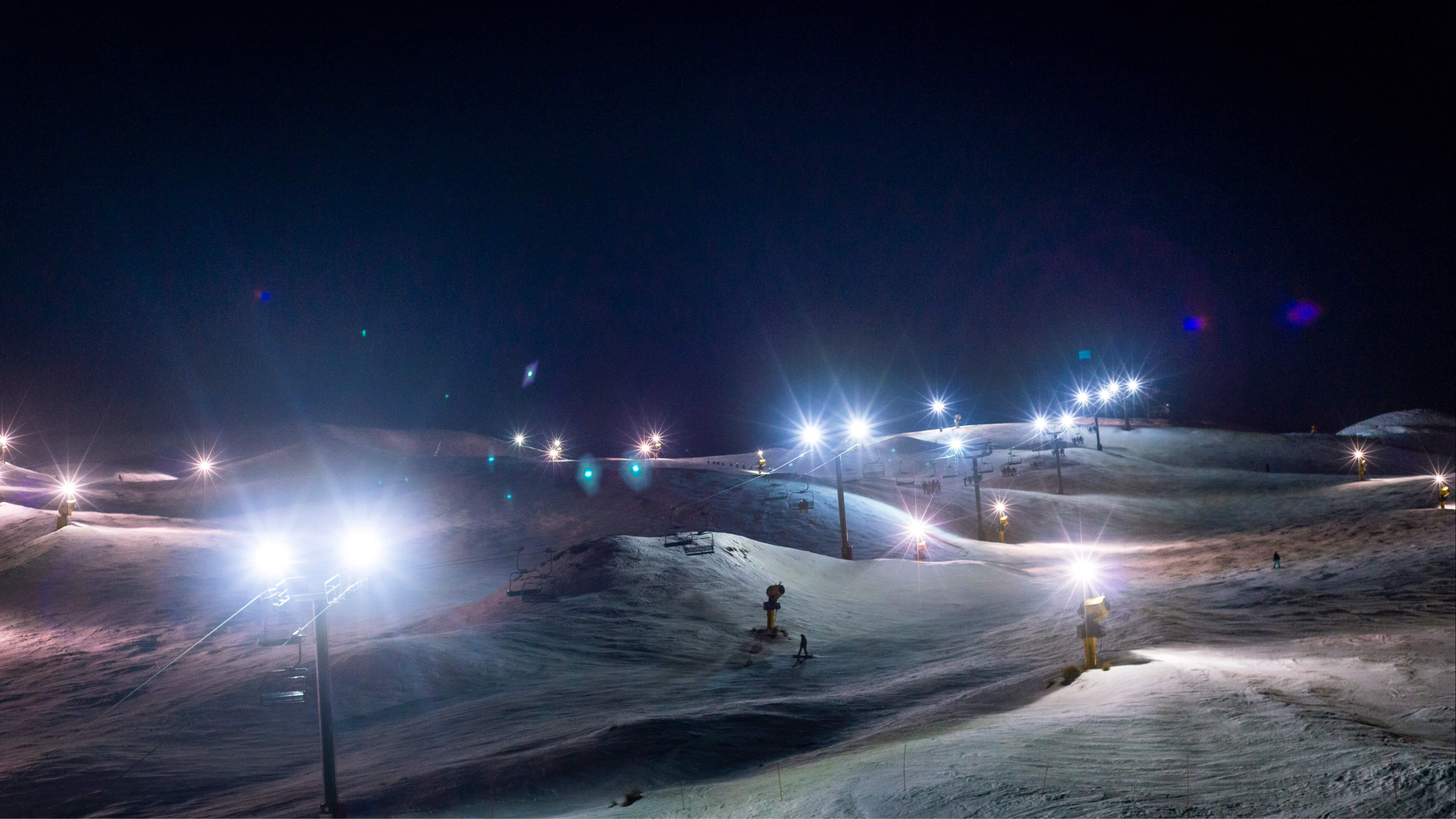 Ski slope at night with bright floodlights and a few skiers on snow-covered hills.