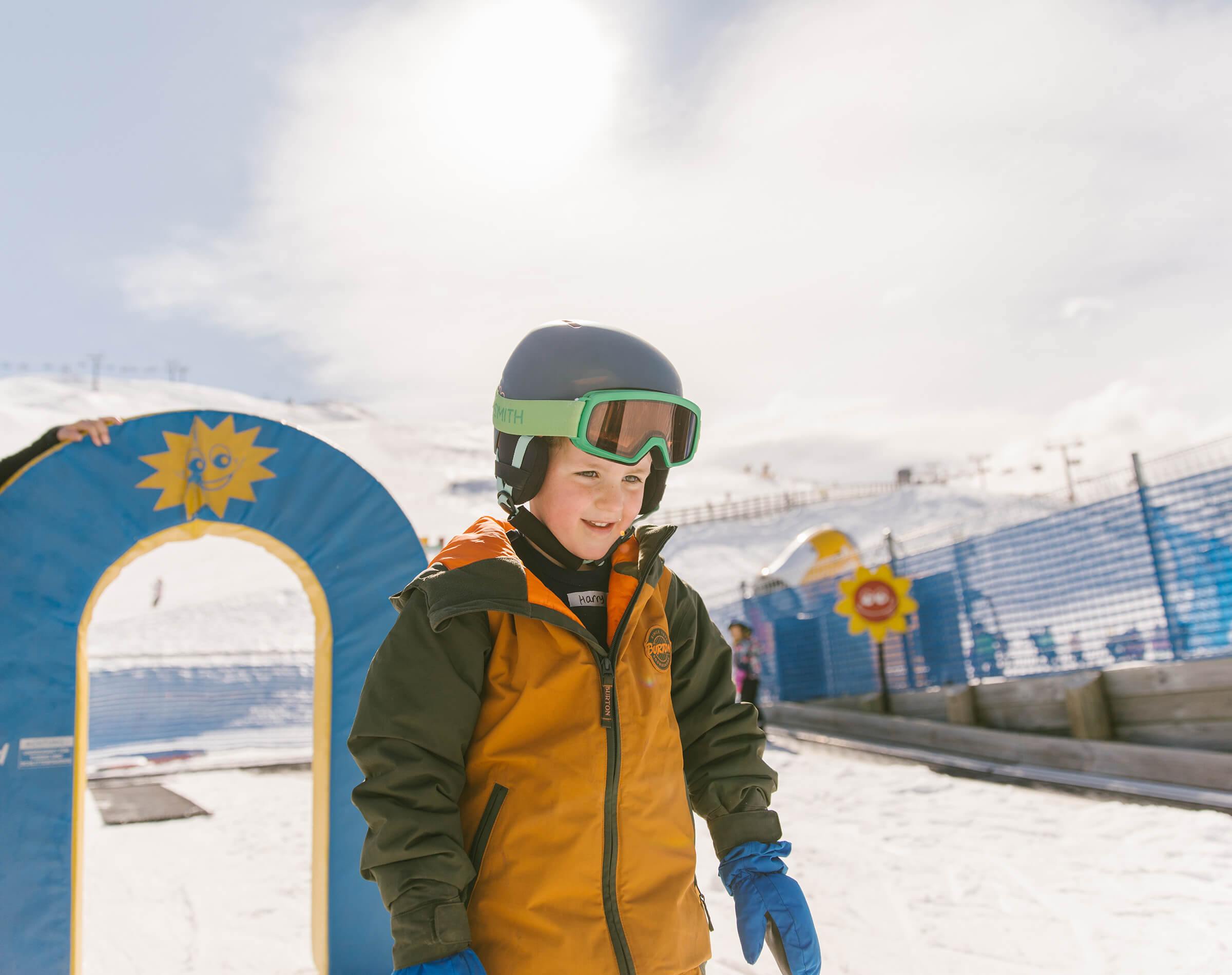 Smiling child in ski gear and helmet at a snowy ski resort on a sunny day.