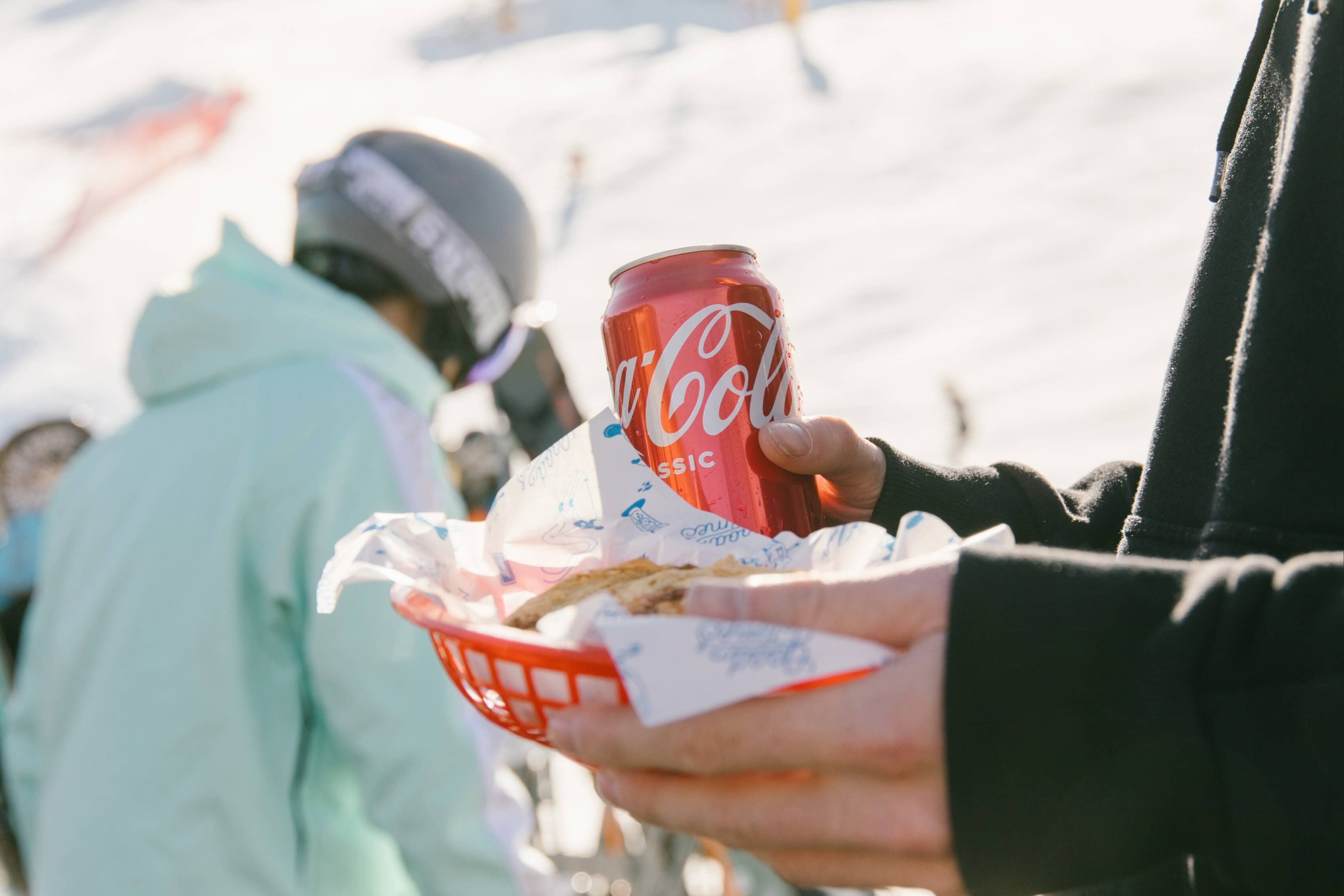 Person holding a can of Coca-Cola and a red basket with food wrapped in paper outdoors.