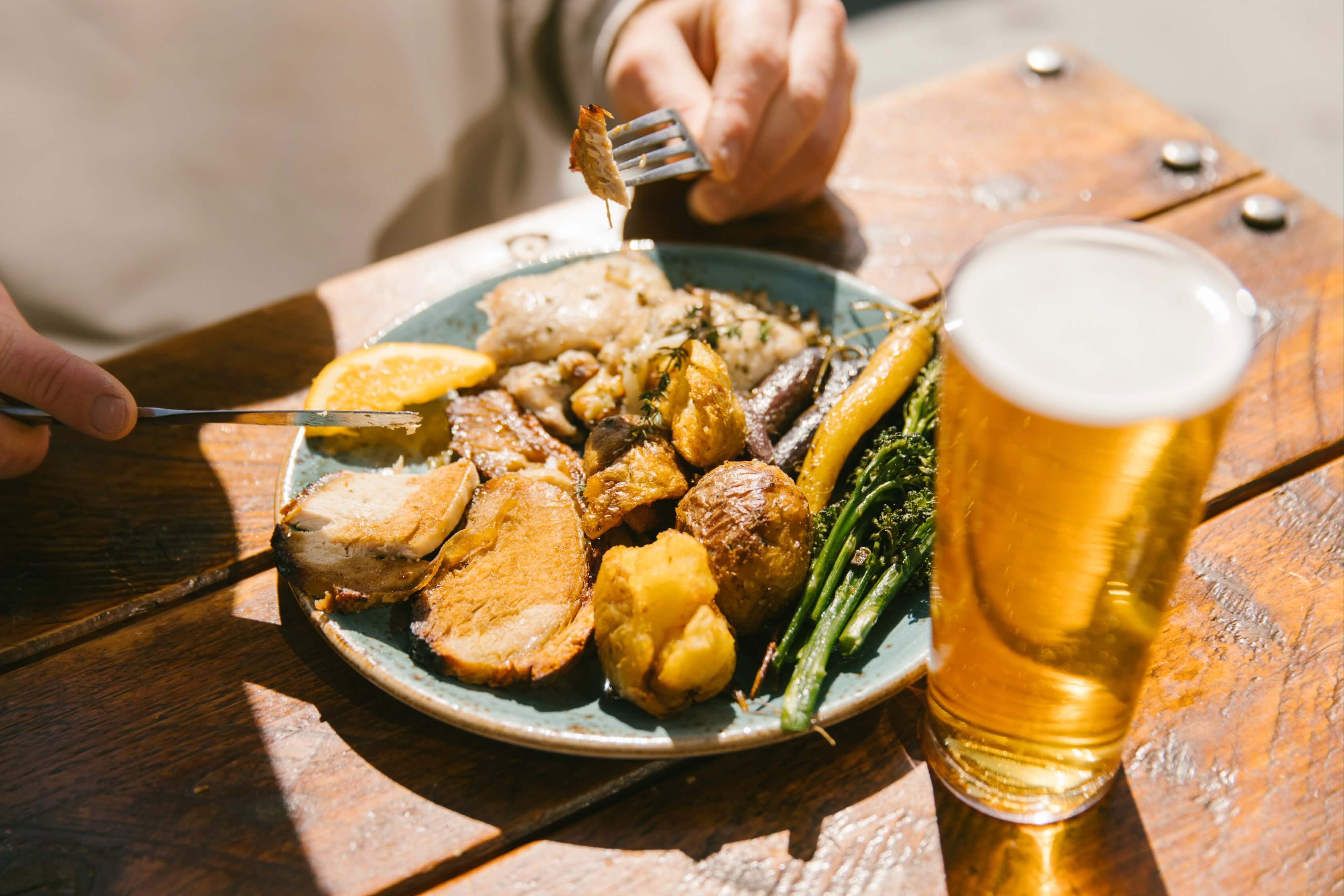 Plate with roasted meat, potatoes, vegetables, and a glass of beer on a wooden table.