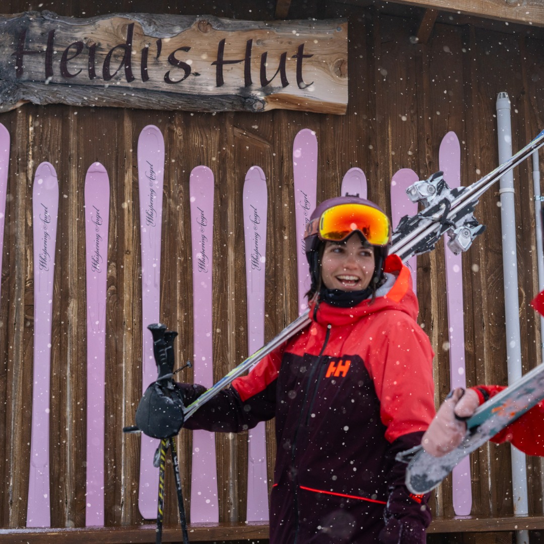 Smiling skier in red jacket holding skis with a wooden sign reading Heidi's Hut behind in snowfall.