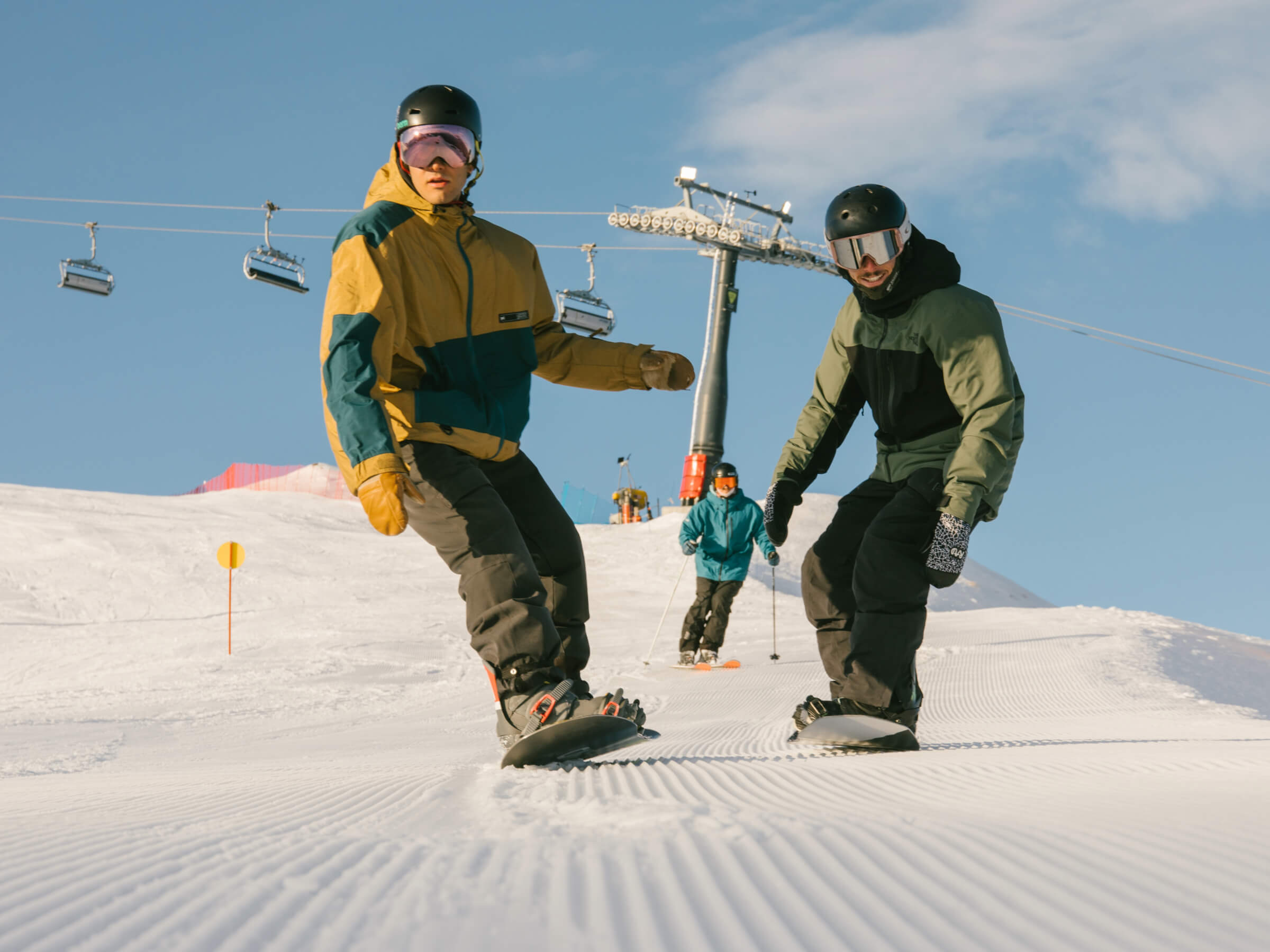 Two snowboarders riding down a snowy slope with a skier behind under a blue sky.