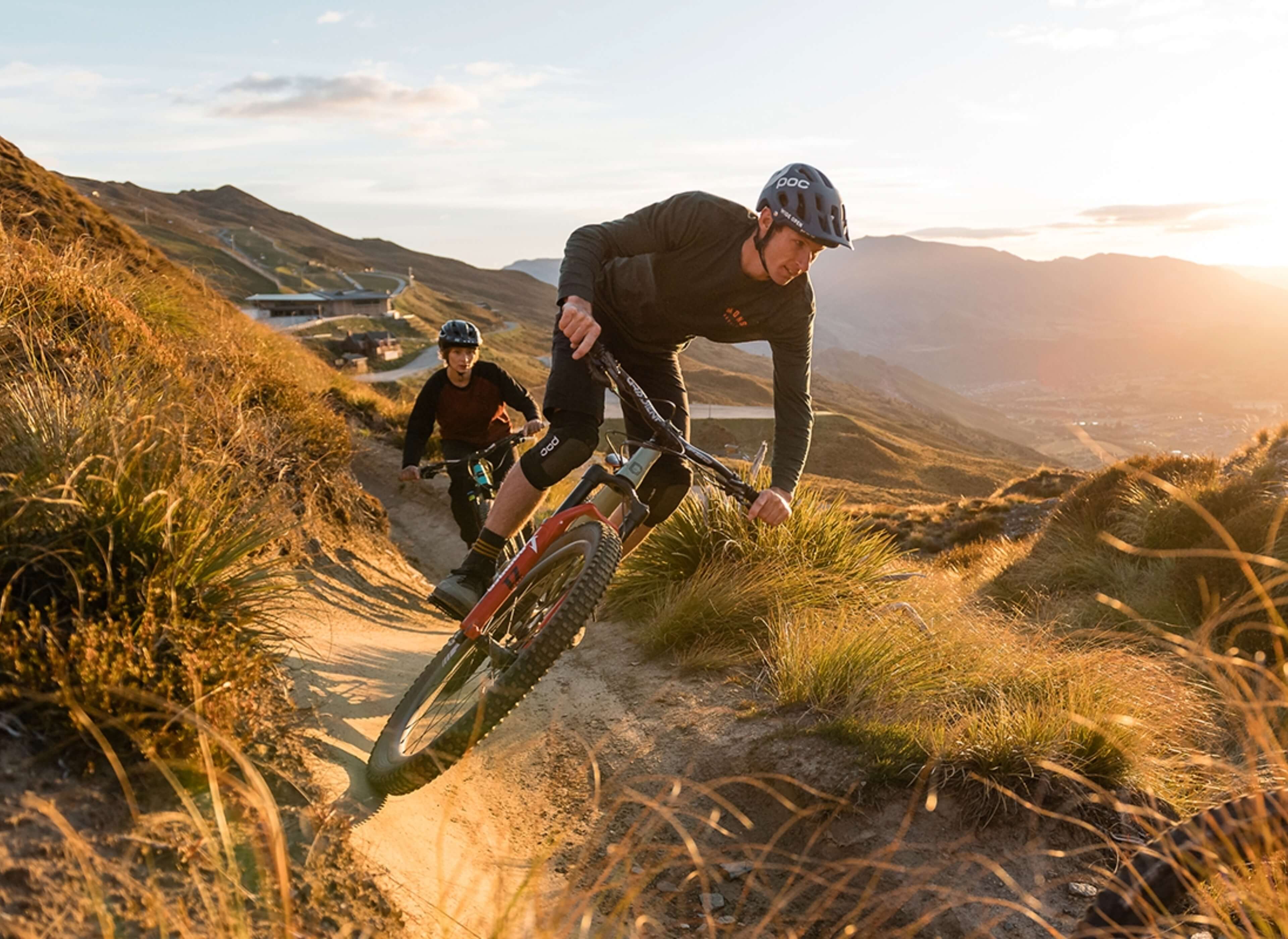 Two mountain bikers wearing helmets riding a dirt trail on grassy hills at sunset.