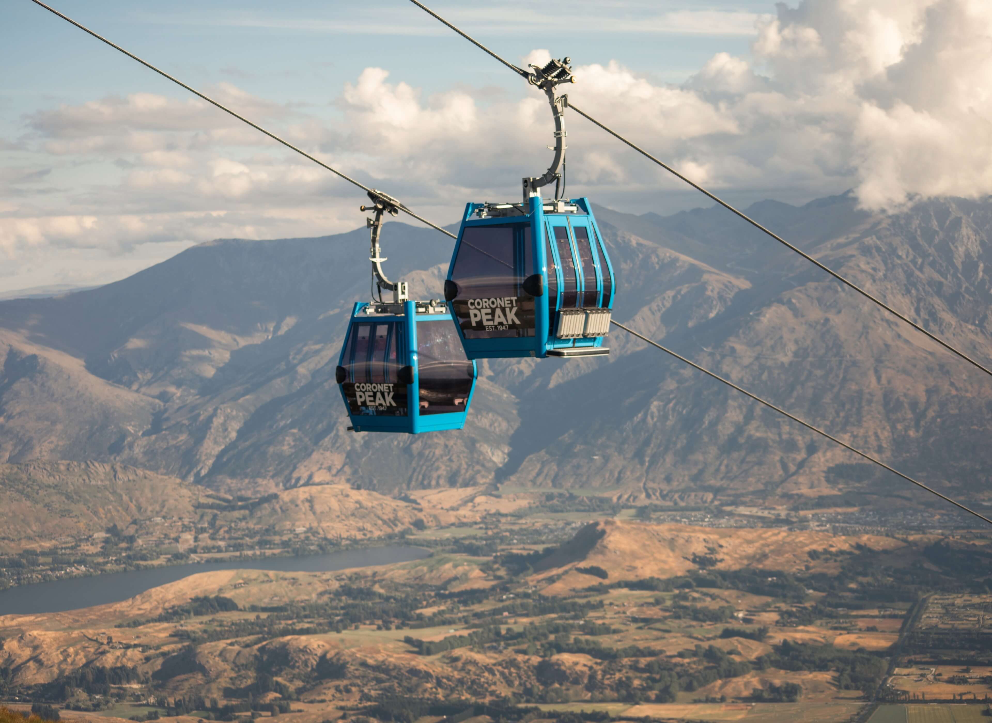 Two blue Coronet Peak gondolas suspended over mountainous terrain with a river and cloudy sky.