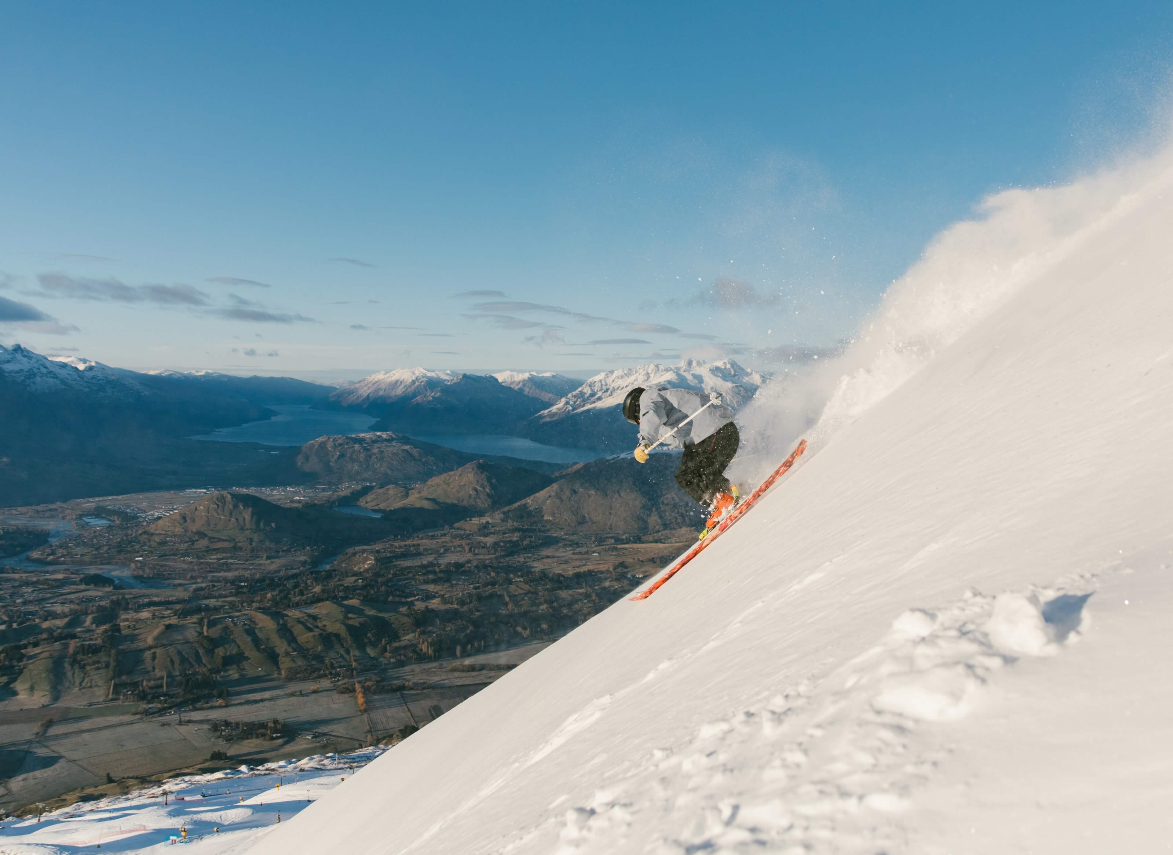 Skier making a sharp turn on a snowy mountain slope with distant mountains and lake below.