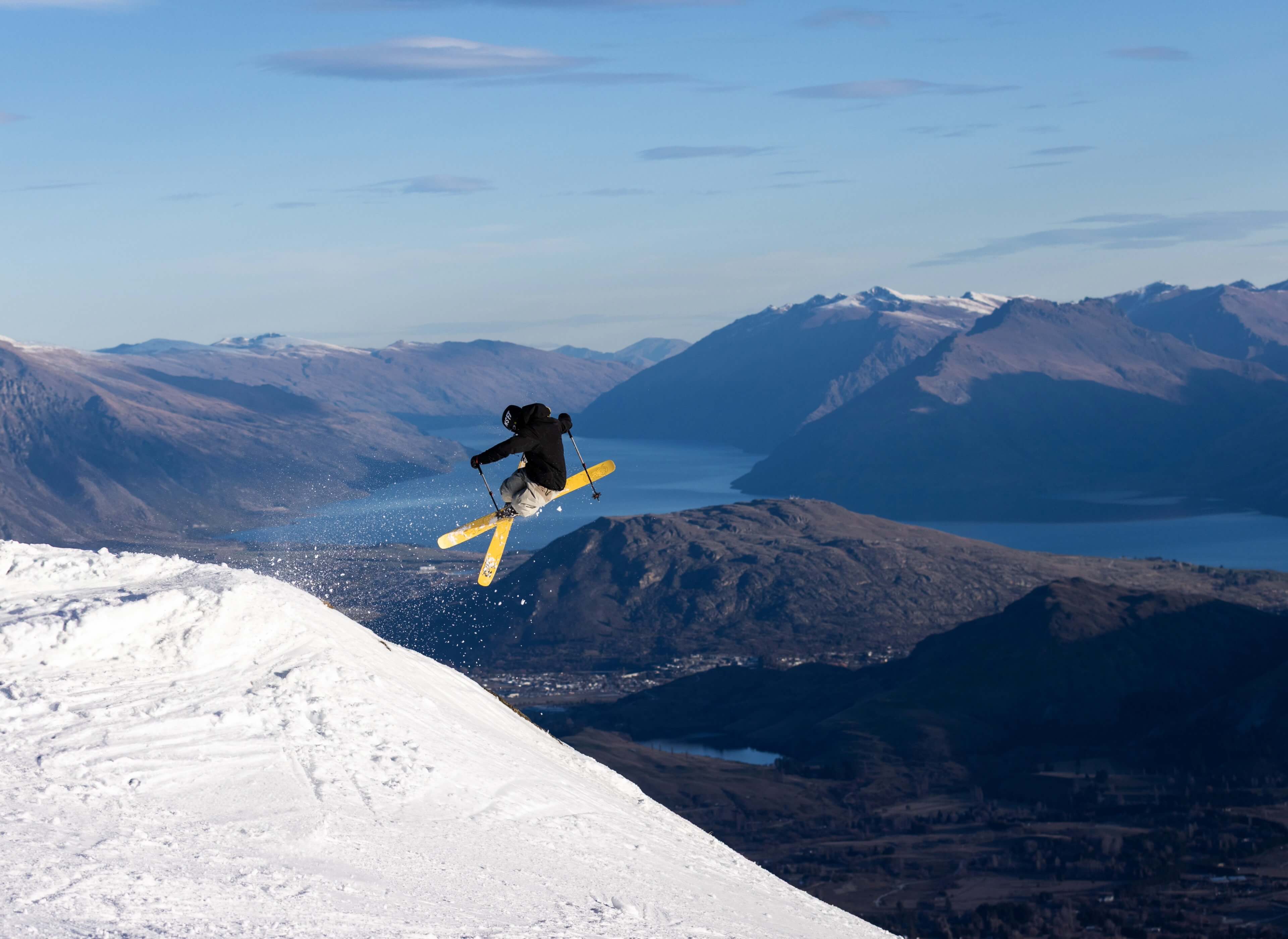 Skier with yellow skis airborne on a snowy slope with mountains and a lake in the background.