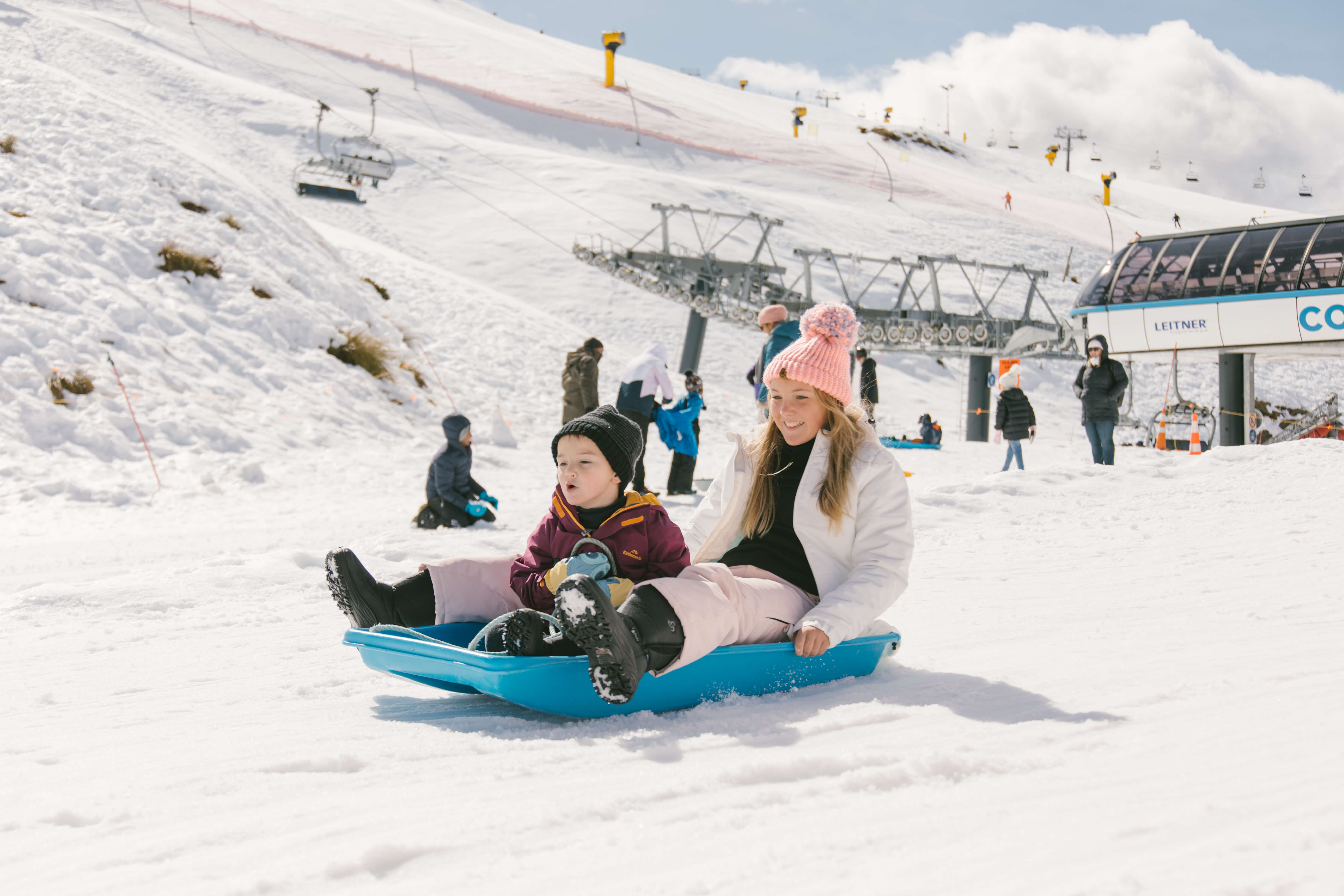 Woman and child sledding down a snowy hill at a ski resort with ski lifts in the background.