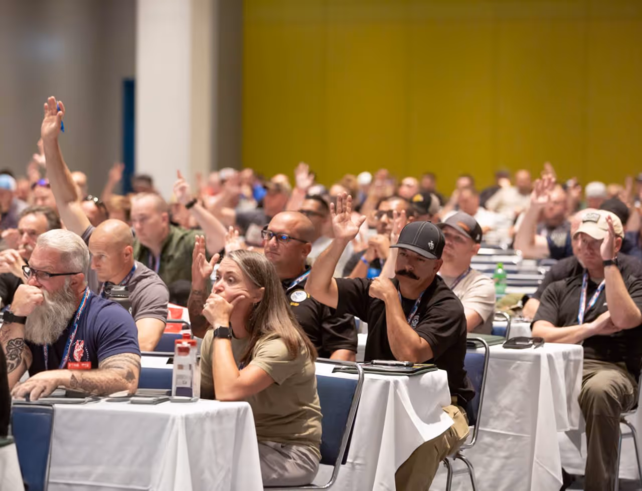 Attendees at a conference raising their hands, seated at tables with white tablecloths.