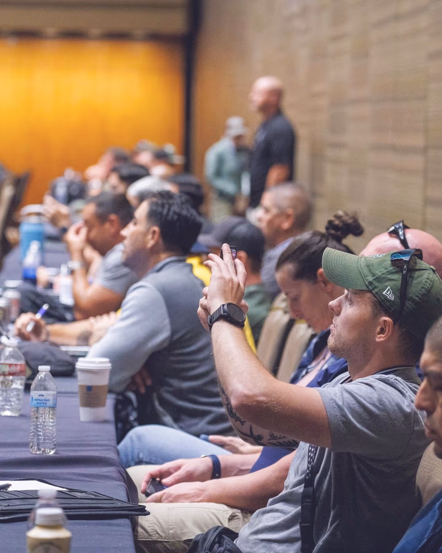 People seated in rows at an indoor event, with one man in a green cap taking a photo with his phone.