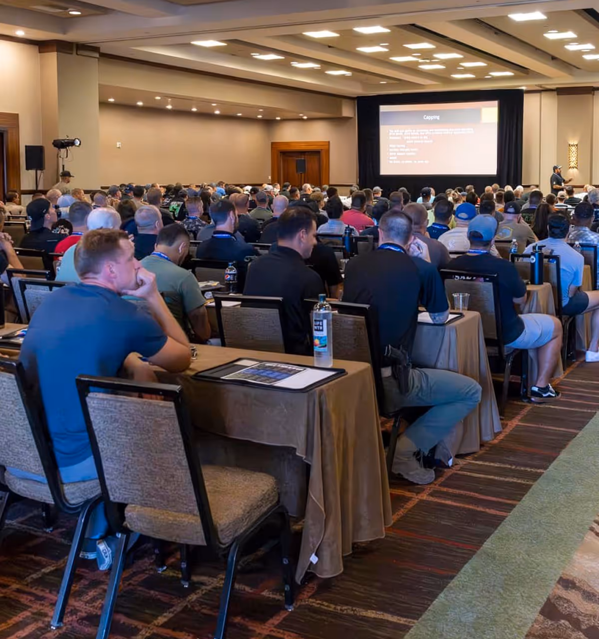 Large group of people seated in a conference room attending a presentation with a speaker at the front near a projection screen.