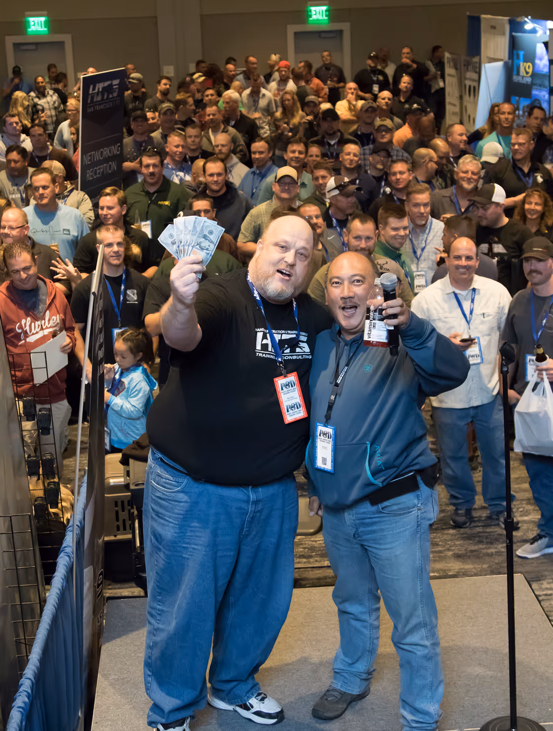Two men smiling at an event, one holding money and the other a drink bottle, with a crowd behind them.