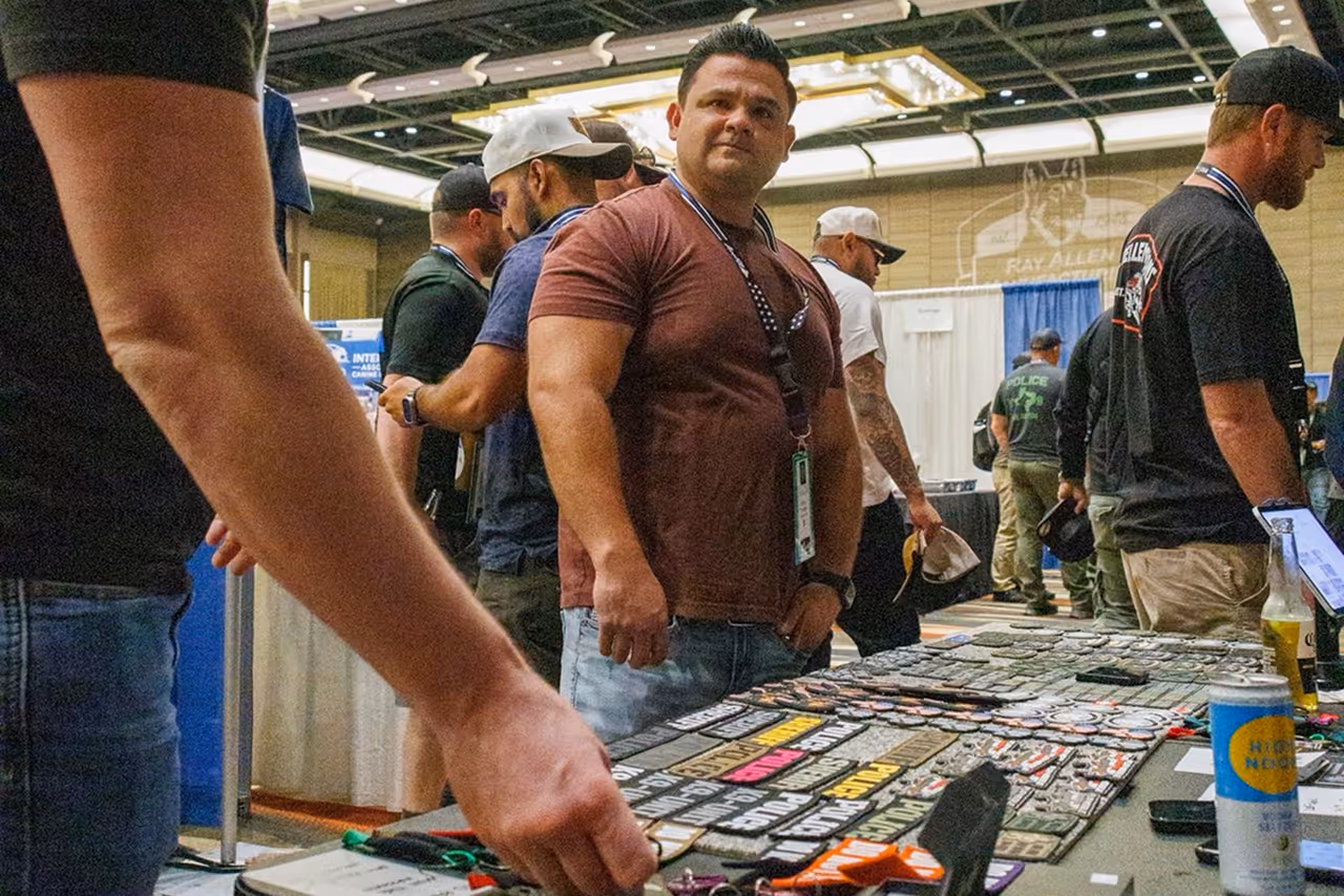 Men browsing patches and gear on tables at a vendor booth in a large indoor conference hall.