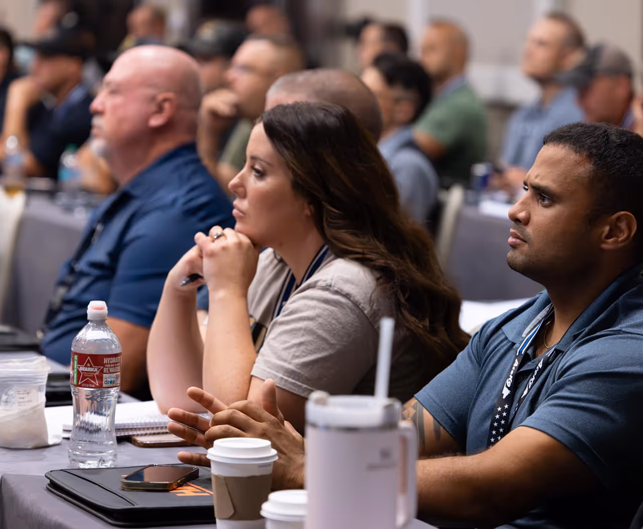 People attentively listening at a conference, sitting at tables with notebooks and drinks.