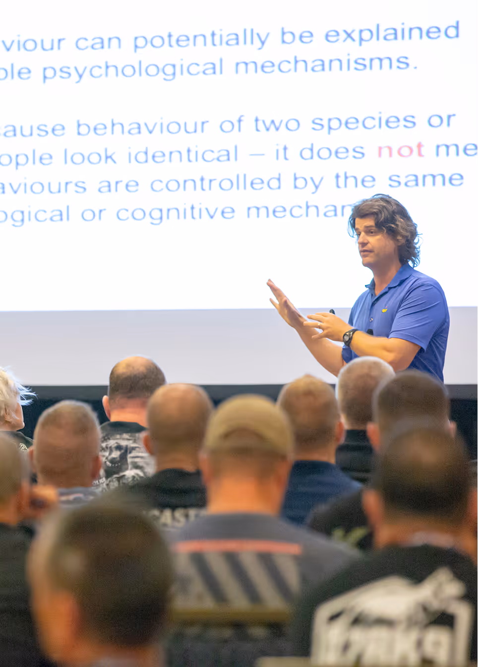Man in blue shirt giving a lecture to seated audience with a large screen of text behind him.