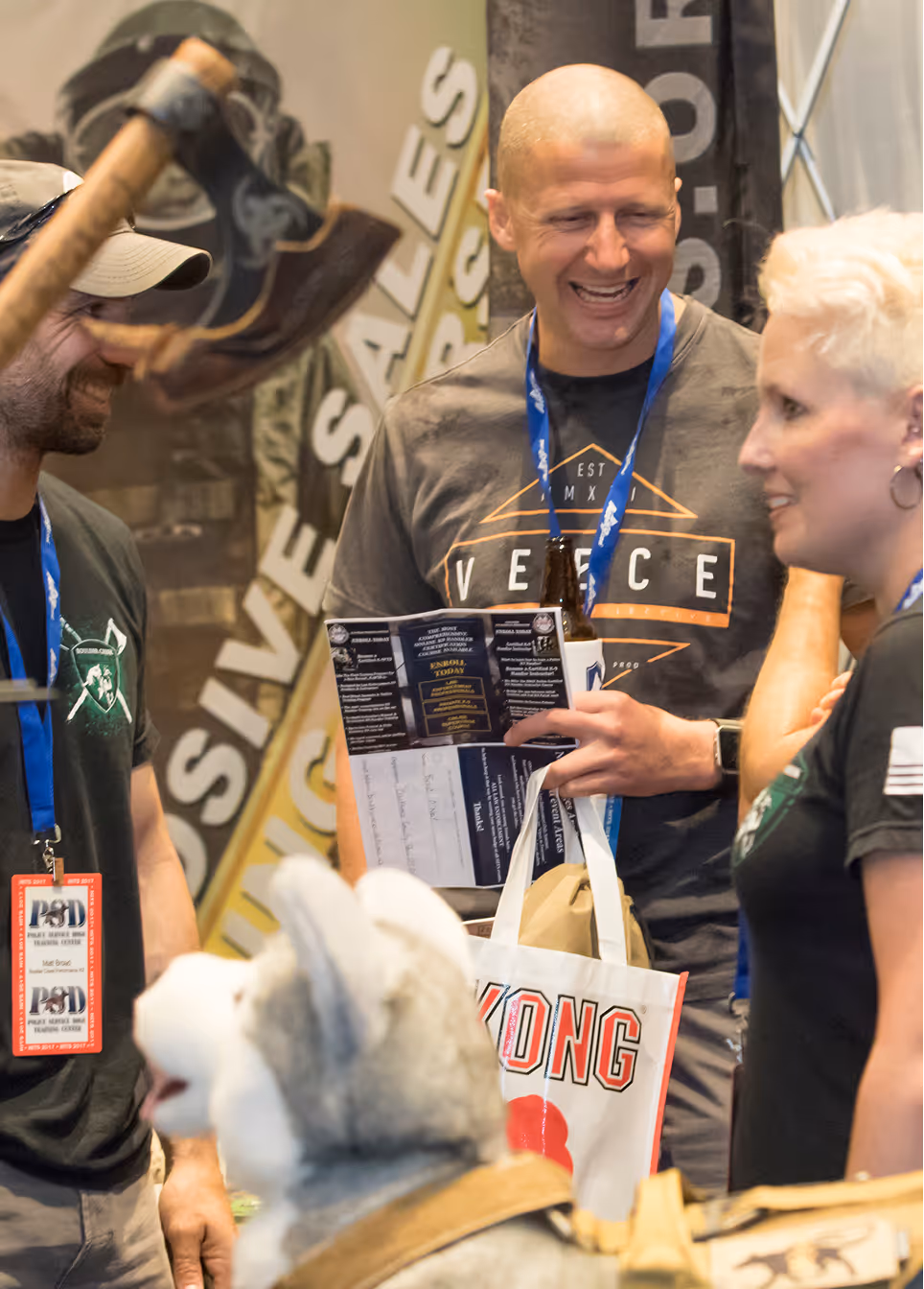 Three people smiling and talking at an indoor event with a dog in the foreground.