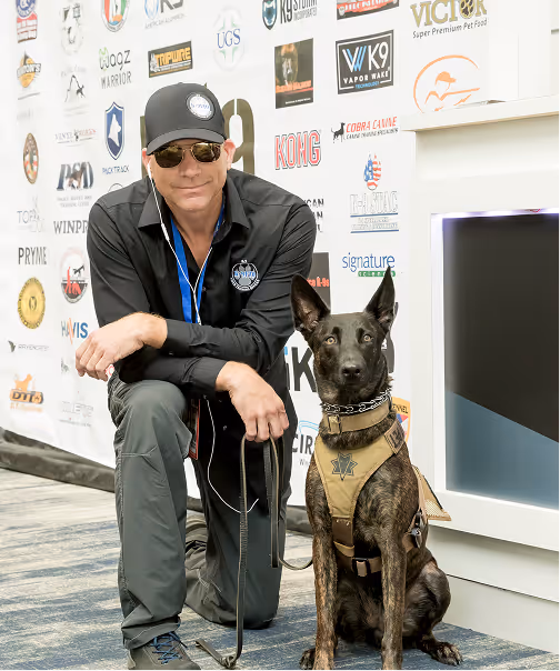 Man in sunglasses kneels beside alert service dog wearing a tan vest indoors.