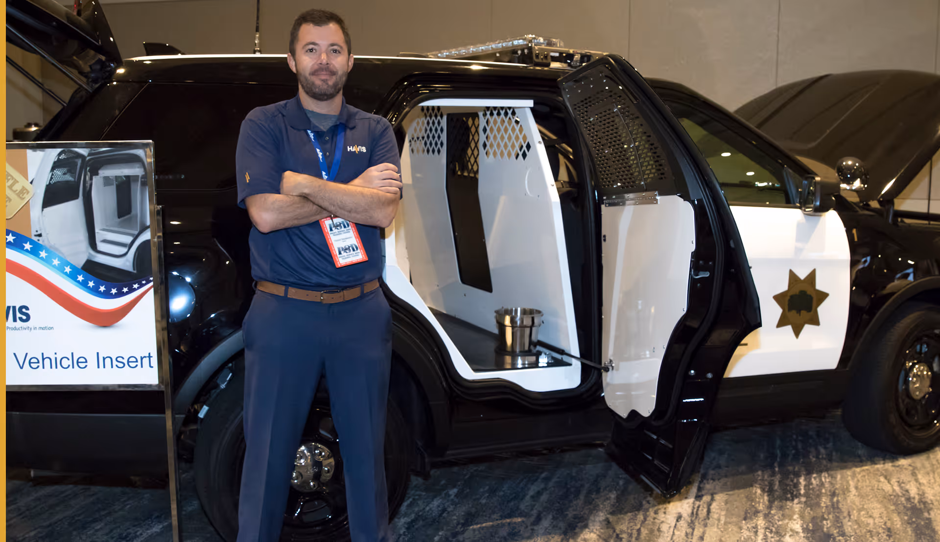 Man stands with arms crossed next to a black and white police car with open rear door showing interior insert.