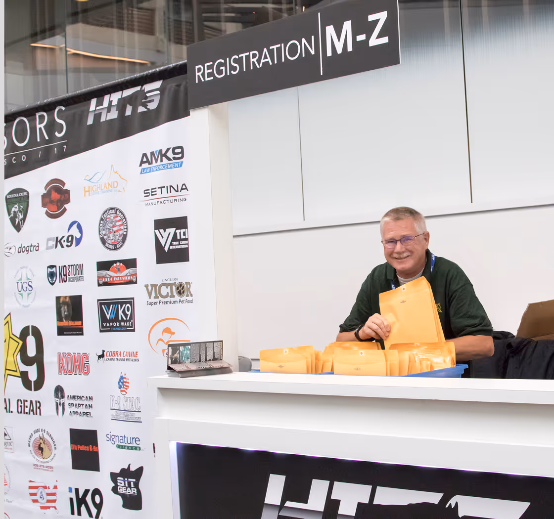 Man wearing glasses smiling behind registration desk with envelope packets under sign M-Z.