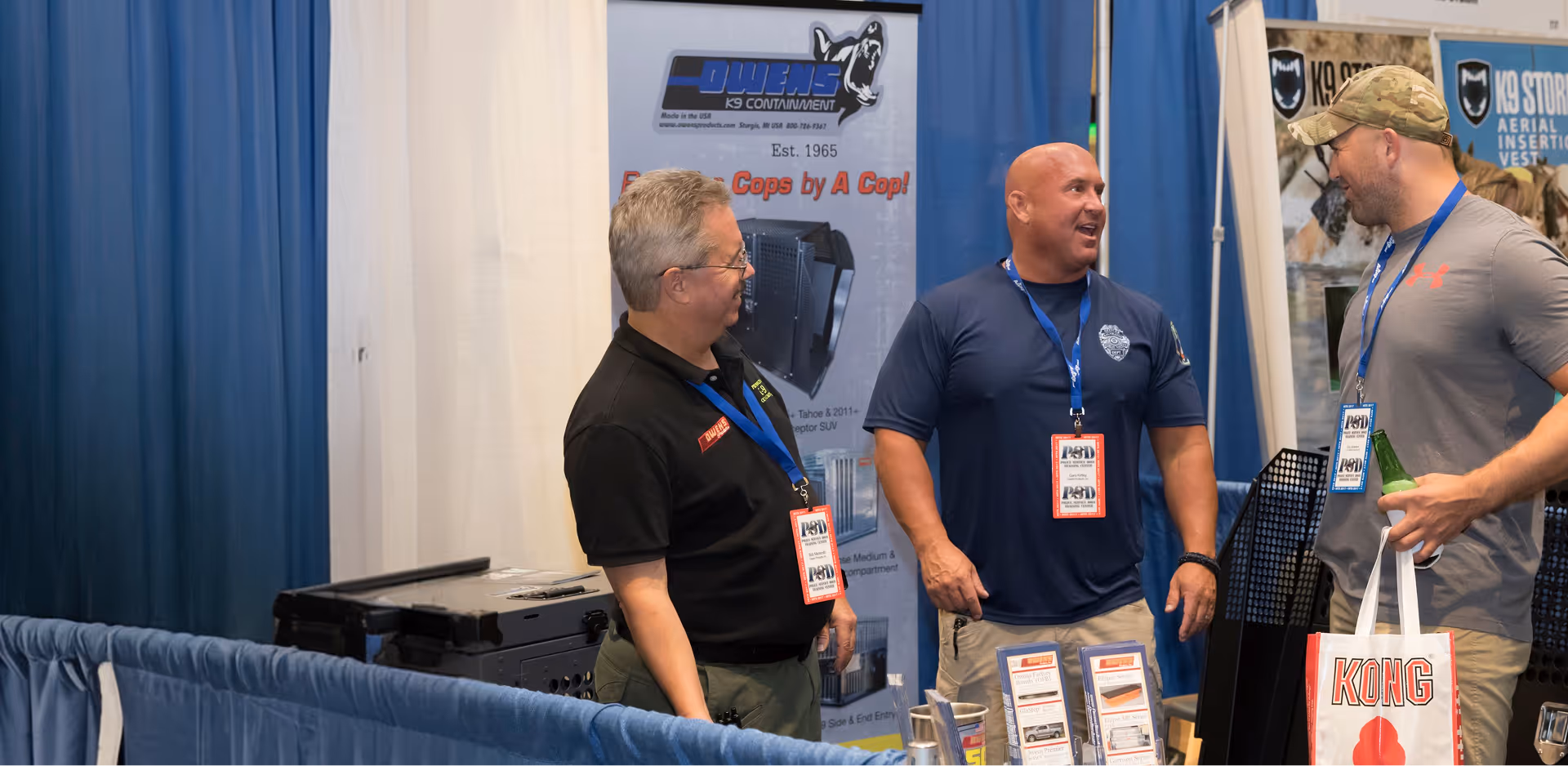 Three men talking at a booth with Owens K9 Containment and K9 Store banners in background.