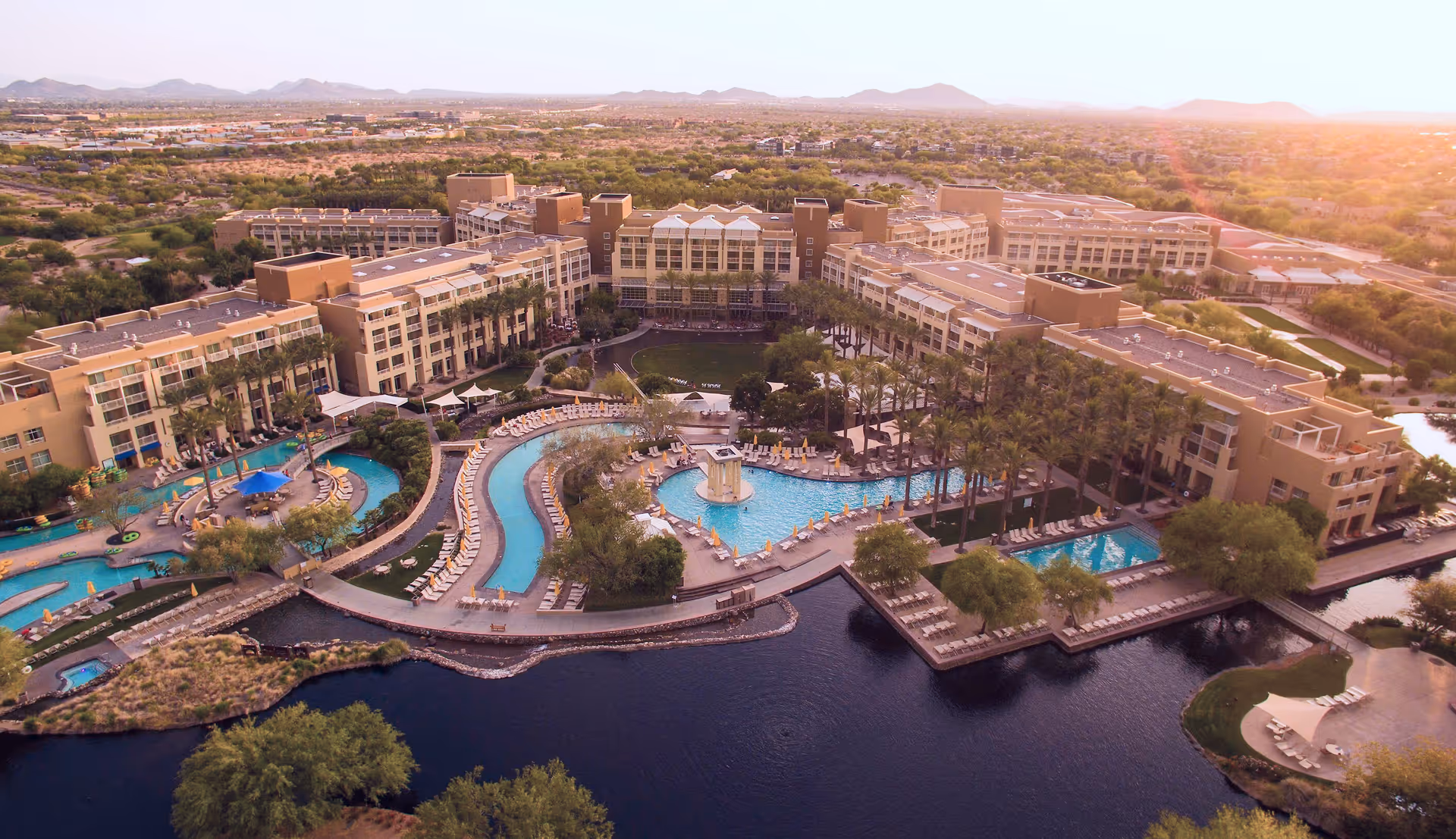Aerial view of a large resort with multiple pools, surrounded by a lake and desert landscape.