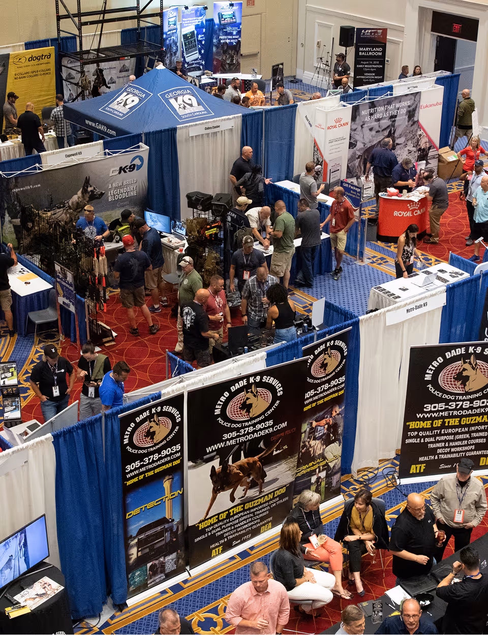 Indoor vendor booths at a dog training expo with displays, banners, and attendees walking and talking.