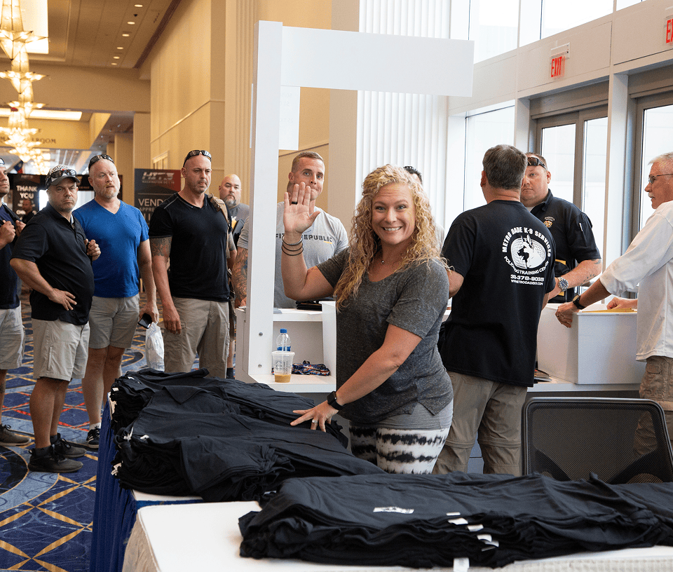 Smiling woman waves behind table with black shirts as men wait in line inside a bright event space.