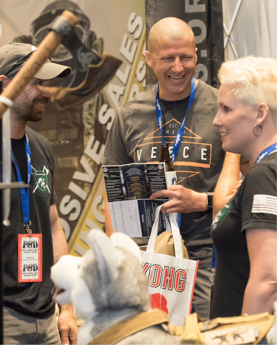 Three people smiling and chatting at an indoor event with lanyards and a service dog in the foreground.