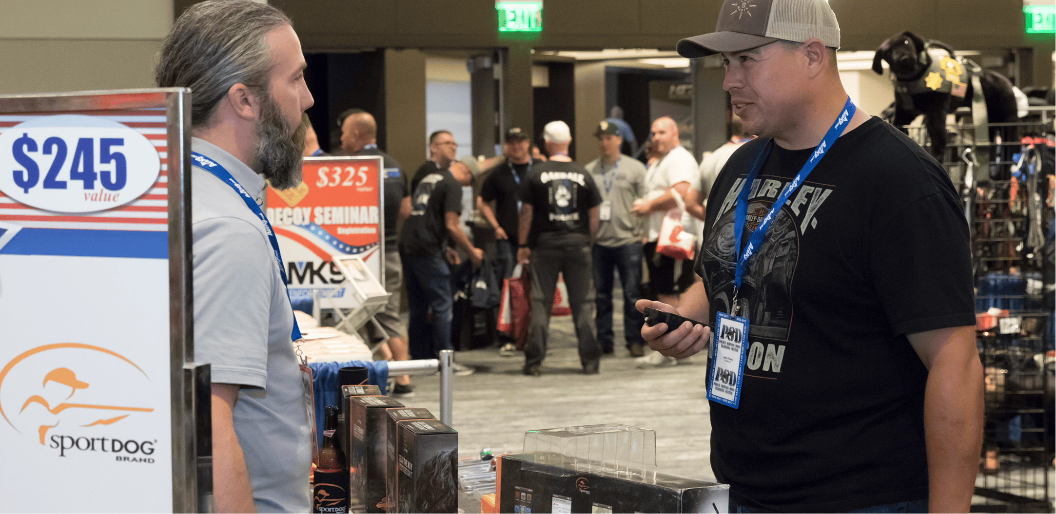 Two men talking at a trade show booth with SportDOG Brand products and displays visible.