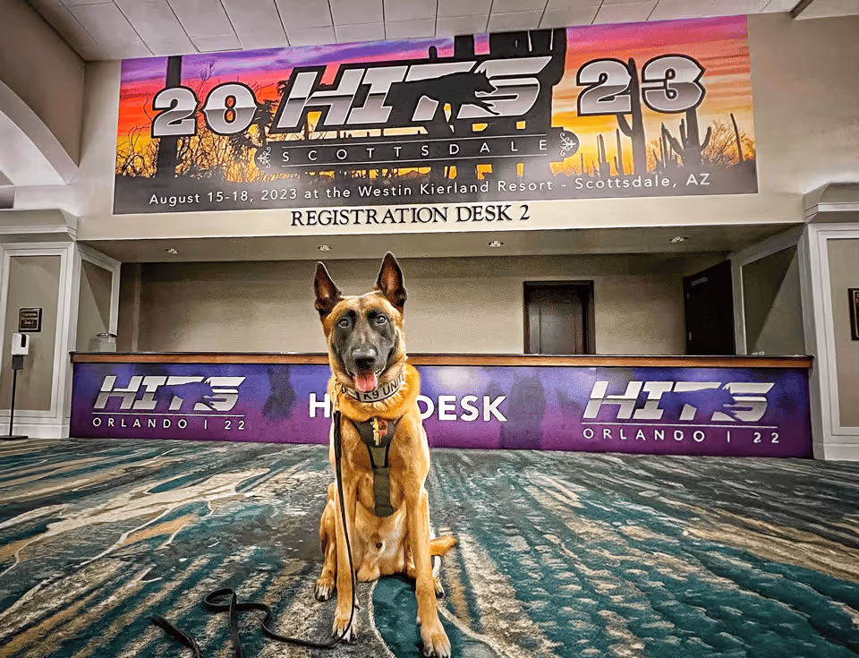 Belgian Malinois K-9 sitting in front of the HITS conference registration desk and Scottsdale 2023 welcome banner

