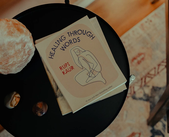 Book titled 'Healing Through Words' by Rupi Kaur on a black round table next to decorative stones and a shell.