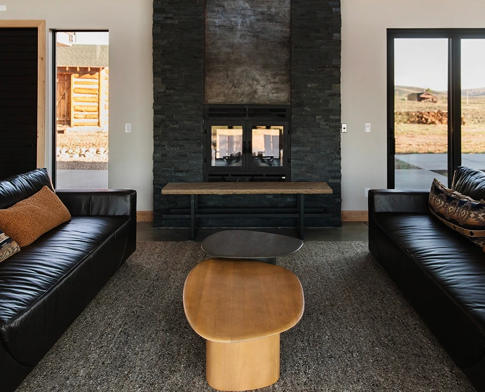 Modern living room with two black leather sofas, a wooden coffee table, a stone fireplace, and large glass doors showing an outdoor view.