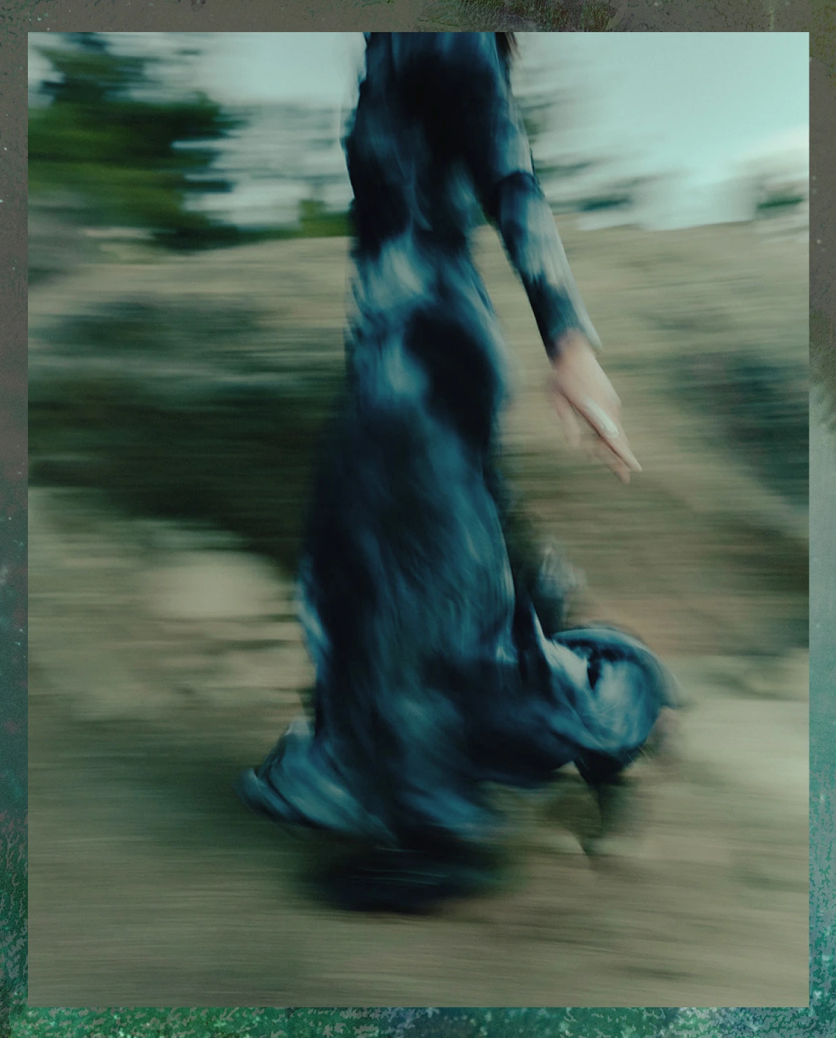 Blurred image of a person in a flowing dark blue dress walking outdoors on a natural ground with greenery in the background.