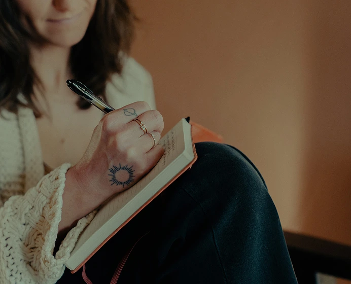 Person with sun tattoo on hand writing in a notebook with a pen, wearing a cream knitted sweater and dark pants.